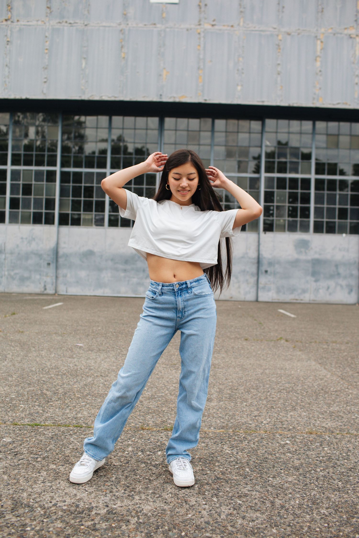 Young woman in white crop top and jeans posing outdoors near industrial building, hands in hair.