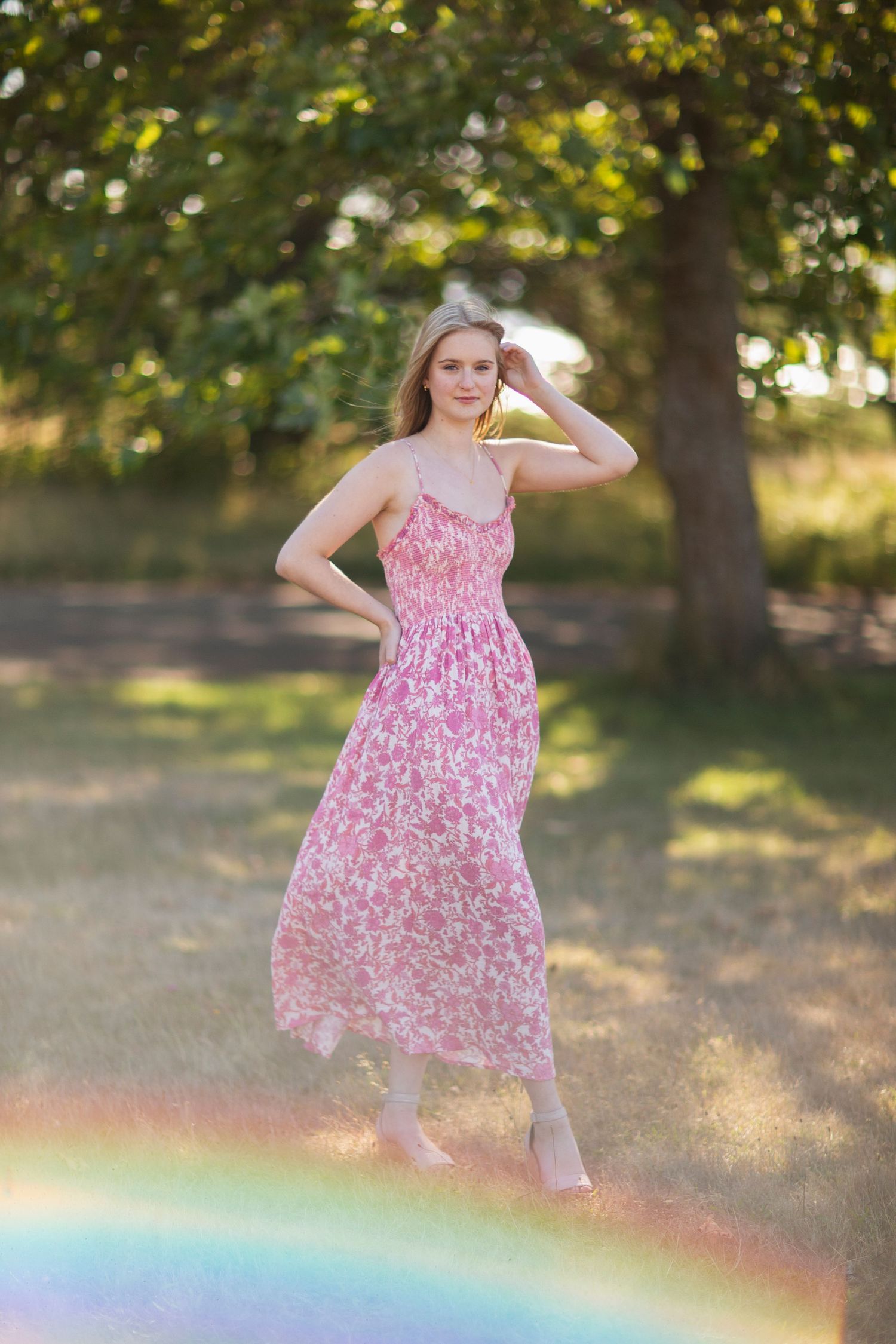 Blonde woman in pink floral maxi dress posing gracefully in golden sunlit field with rainbow.