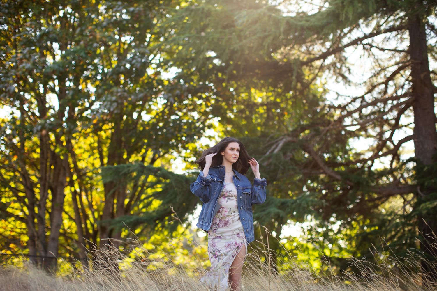 Young woman in floral dress and denim jacket standing in golden sunlit forest clearing.