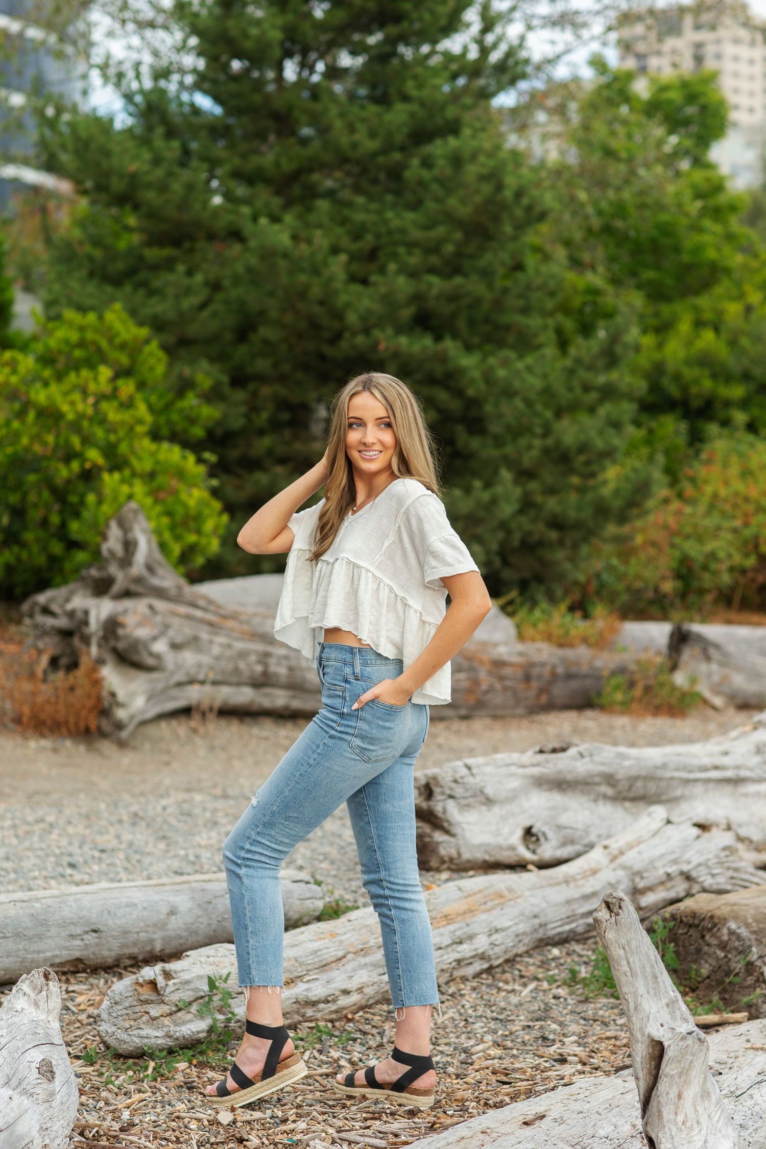 Young woman in white crop top and blue jeans posing on driftwood logs in a natural outdoor setting.