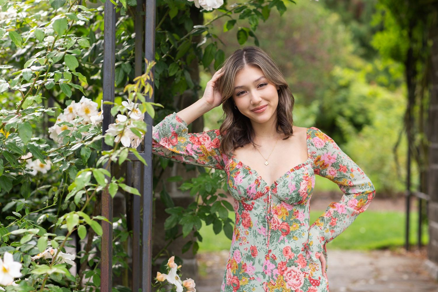 Young woman in a floral long-sleeve dress posing beside white roses in a lush garden setting.