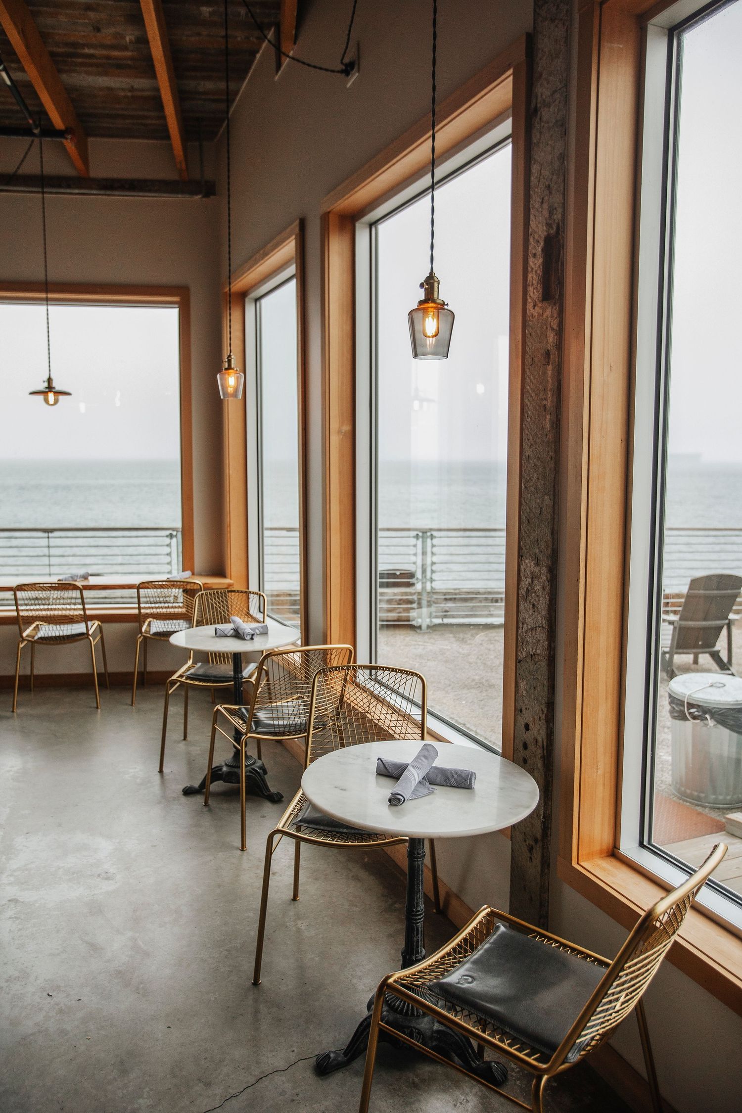 A bright waterfront cafe interior with wooden tables, rattan chairs, pendant lights, and ocean views.