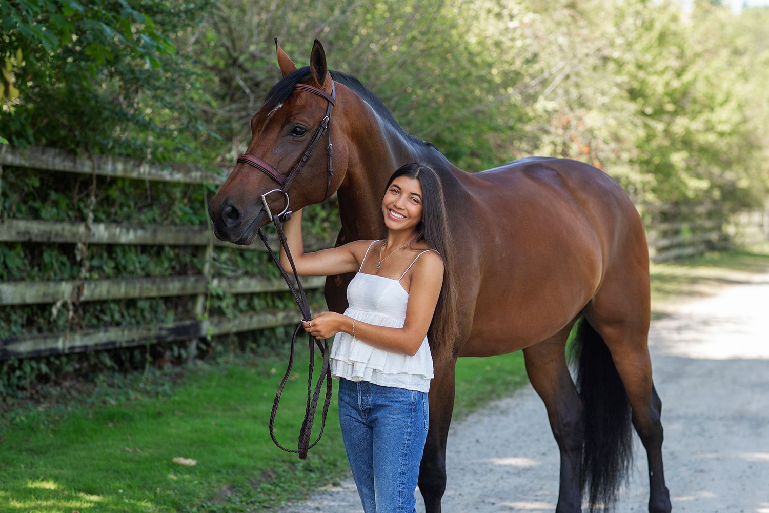 Young woman in white top and jeans smiling beside a chestnut brown horse on a sunny tree-lined path.