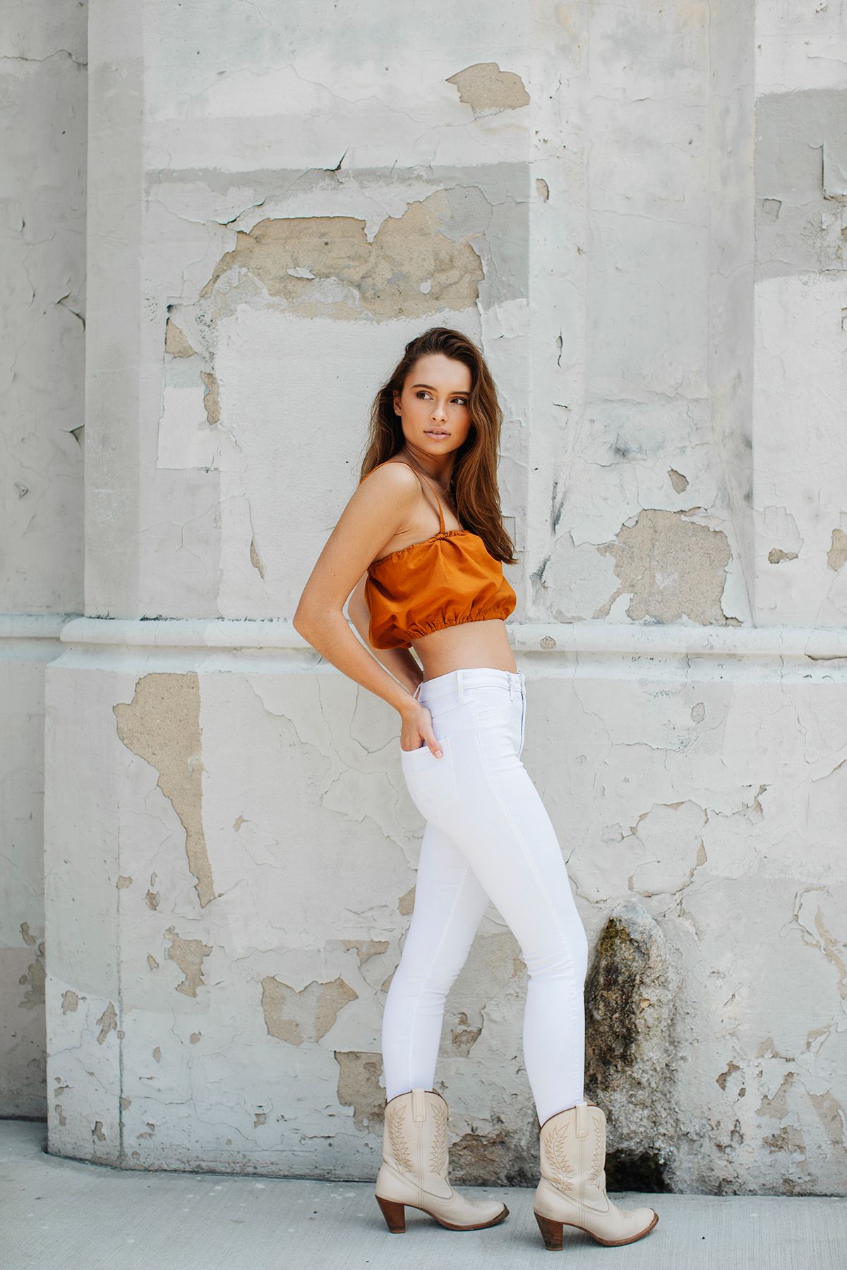 Woman in orange crop top and white jeans poses against distressed white wall with peeling paint.