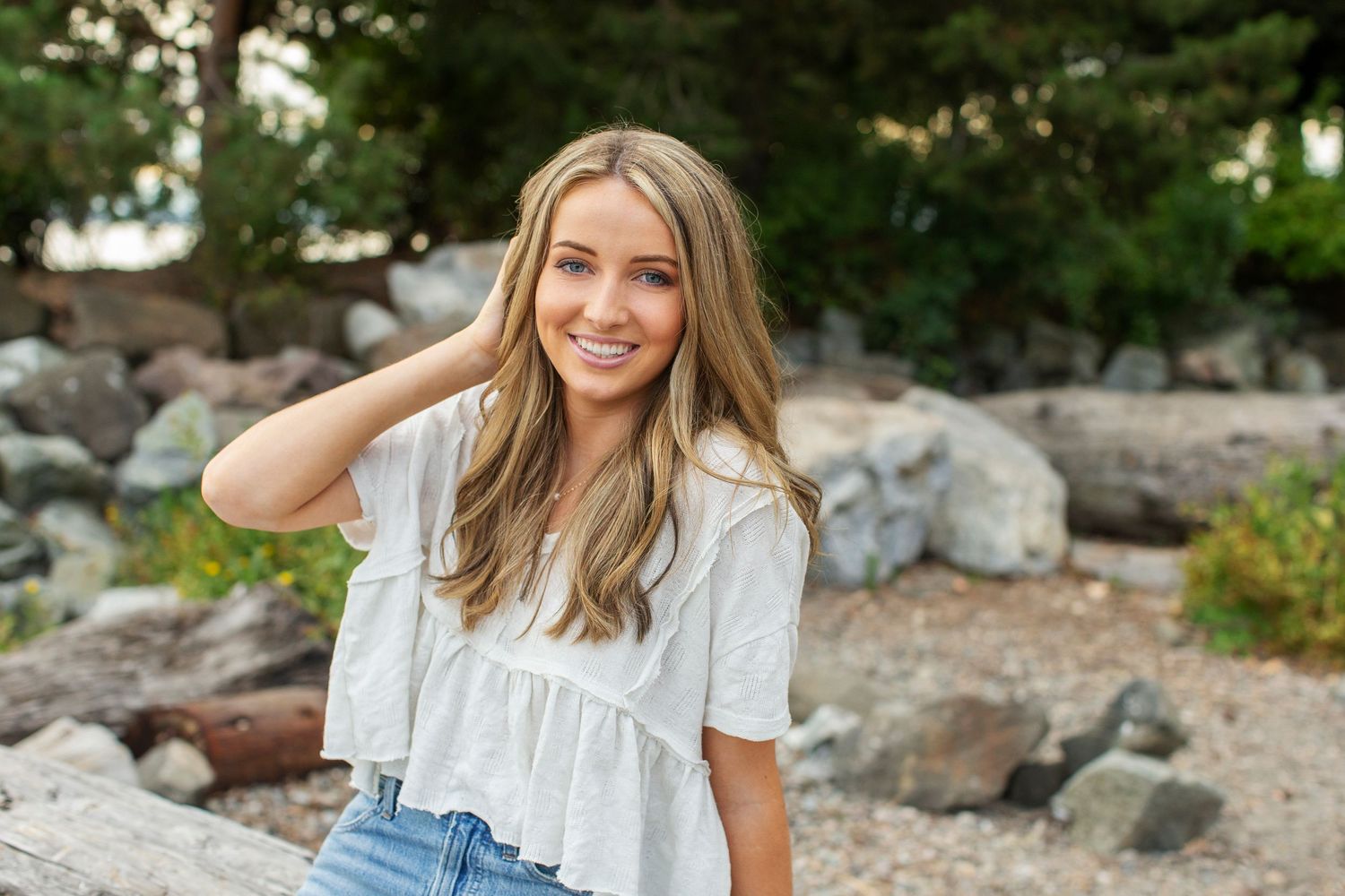Smiling young woman in white ruffled top with hand in hair near rocky background.