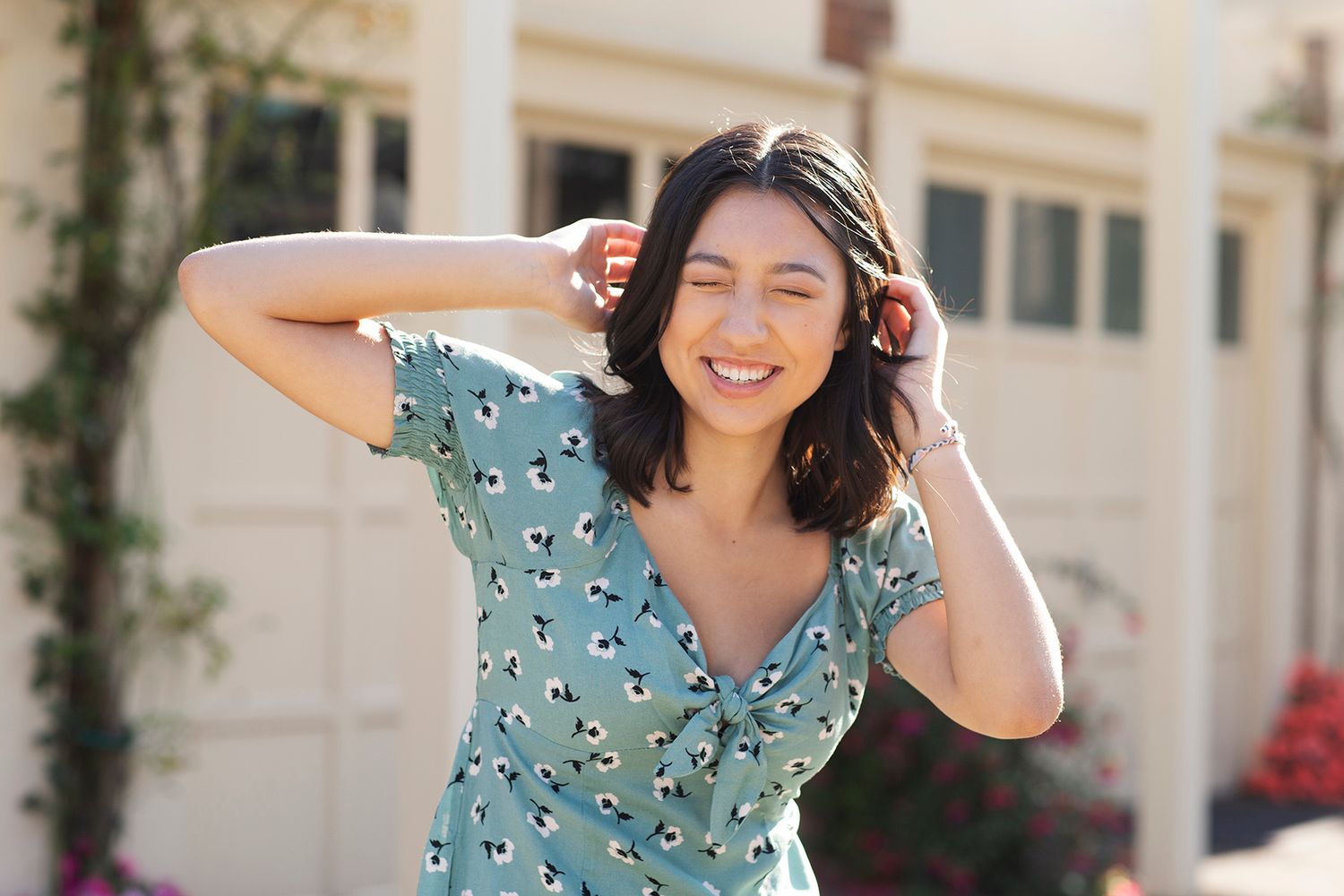 Smiling young woman in a floral teal wrap dress posing outdoors in sunlight with hands in her hair.