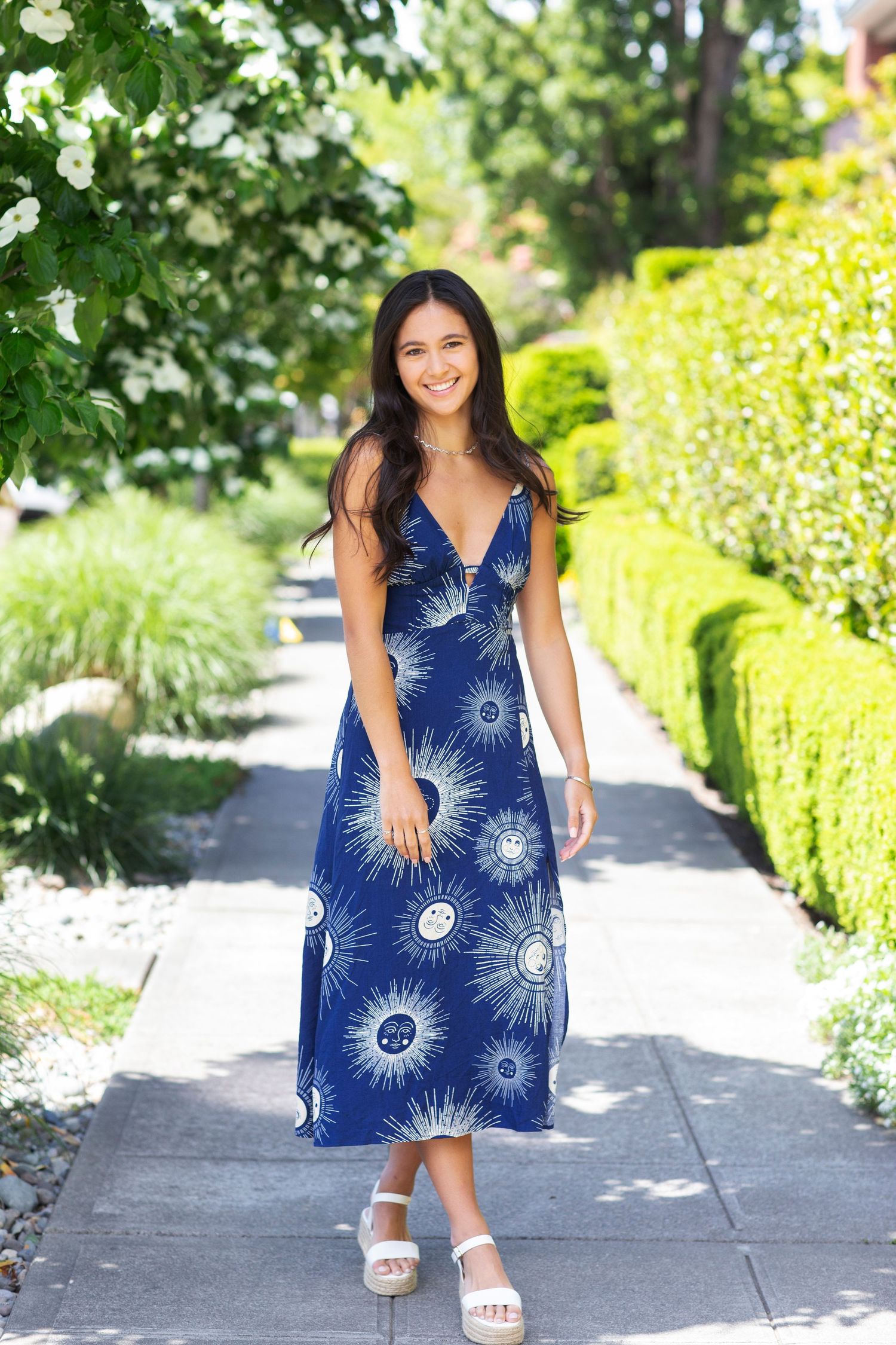 Woman in navy blue sun print midi dress walking on a tree-lined sidewalk wearing white wedge sandals.