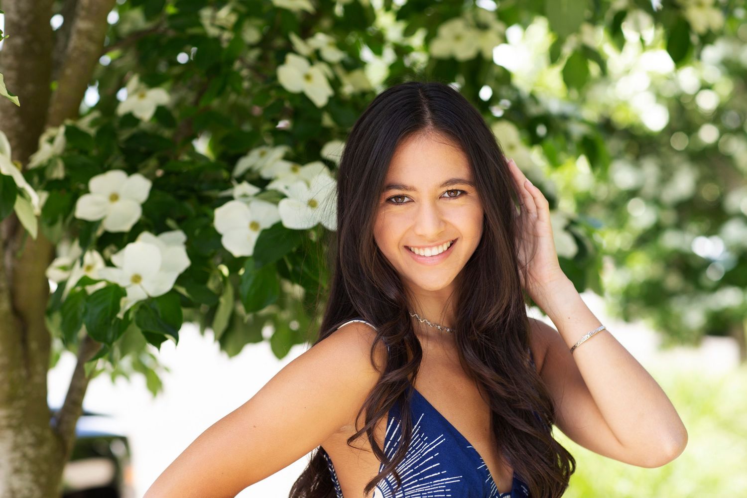 Smiling brunette woman in blue dress posing near white flowering tree outdoors.