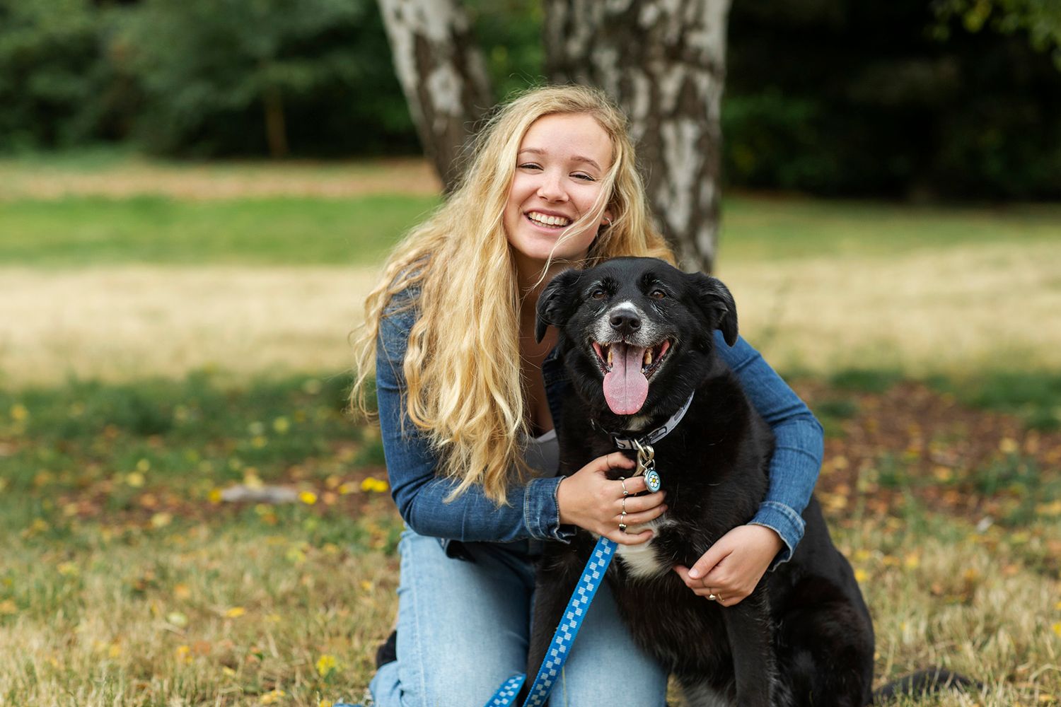 Smiling blonde girl hugging a happy black dog on a leash in a green park setting.