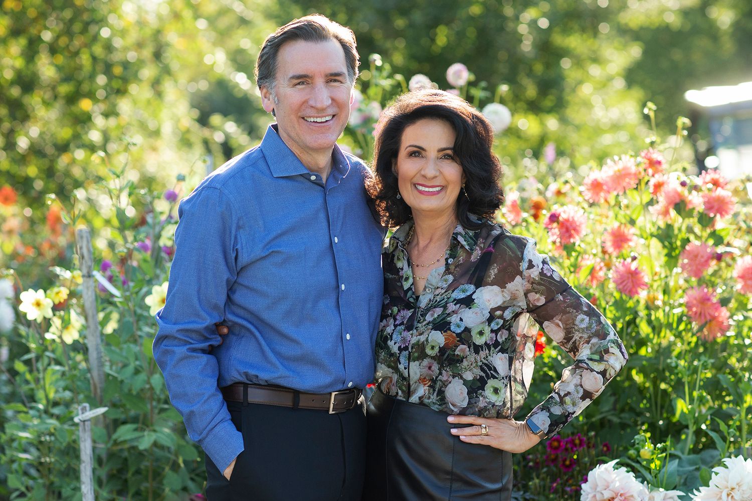 A smiling couple poses together in a vibrant garden filled with colorful dahlias and flowers in bloom.