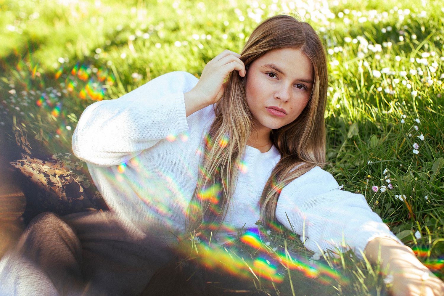 Young woman with long hair sits in green grass field wearing white top, surrounded by rainbow light flares.