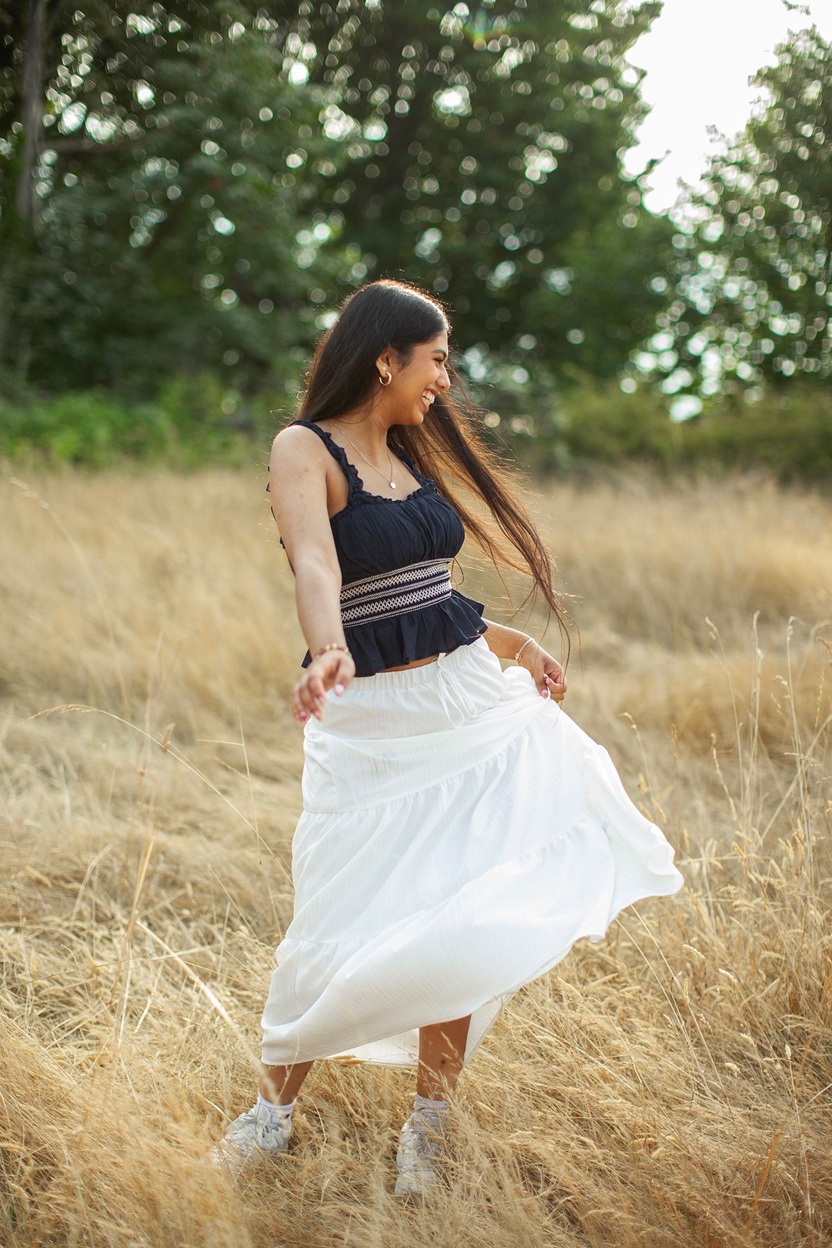 Joyful young woman in black crop top and white maxi skirt dancing in dry grass field.