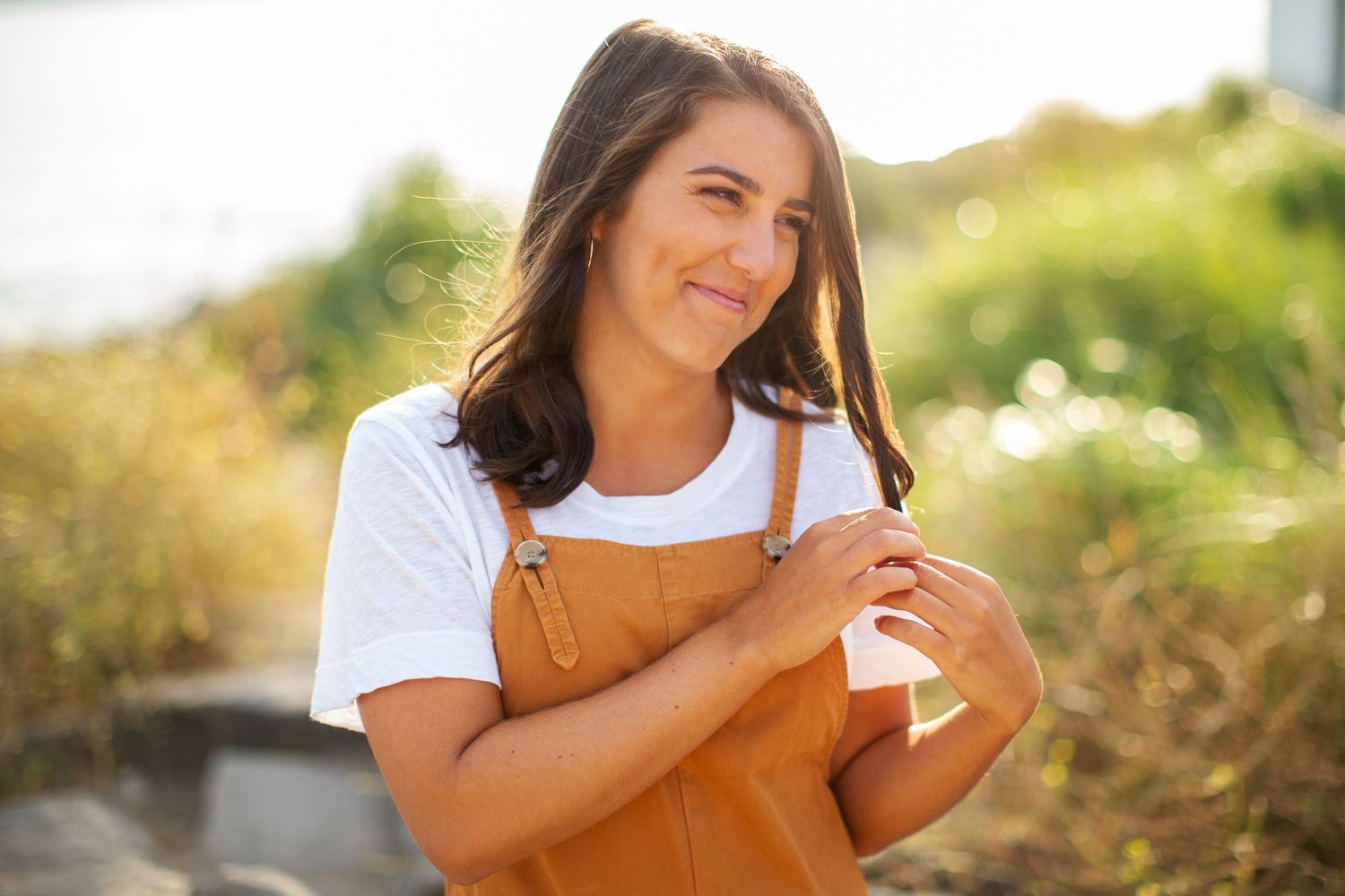 Smiling brunette woman holding hair outdoors in golden sunlight wearing a white t-shirt.