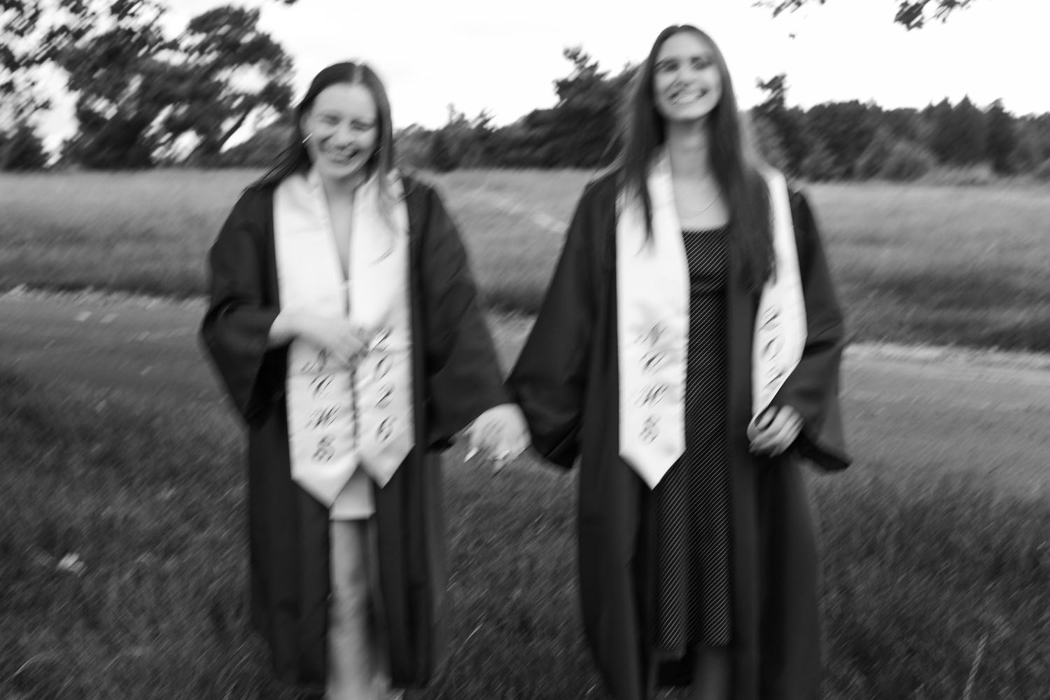 Two smiling graduates in black gowns and white stoles pose outdoors in a blurry black and white photo.