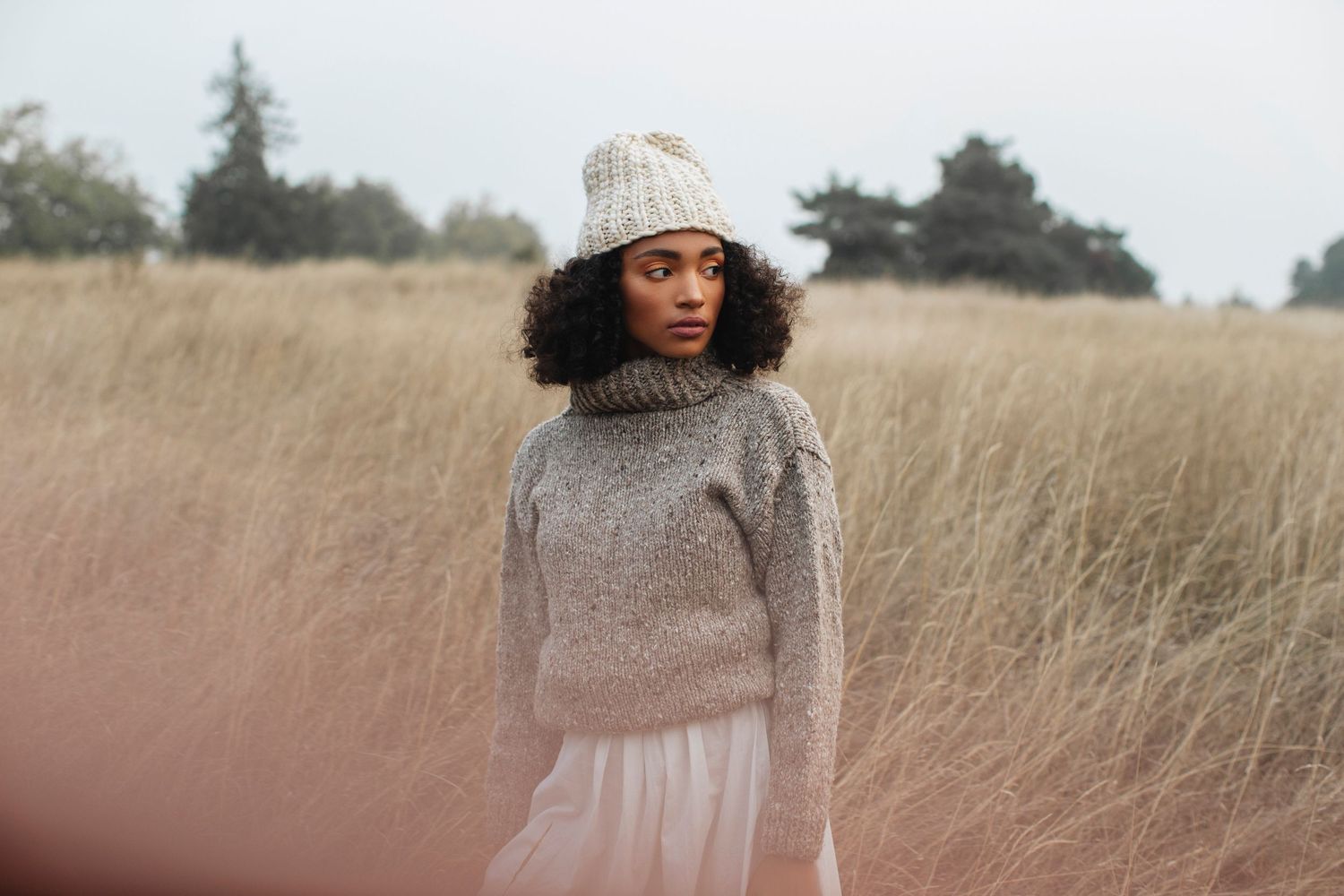 Young Black woman in knit sweater and beanie hat standing in tall golden grass field on overcast day.