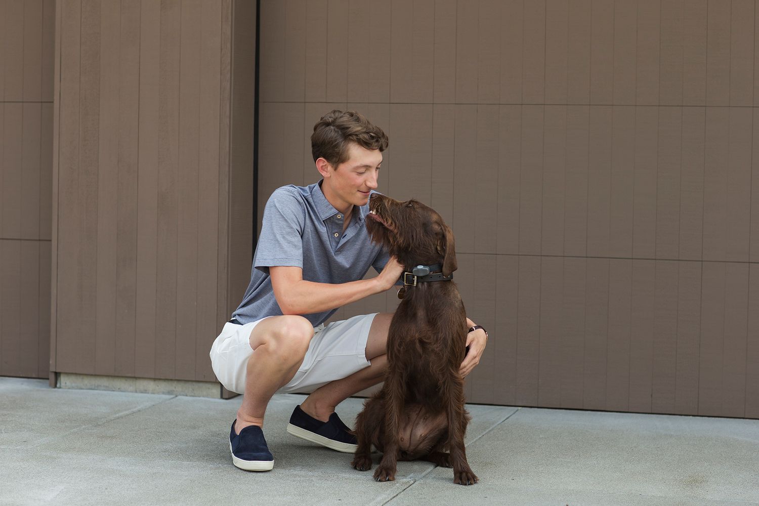 Young man crouching outdoors petting a chocolate brown Cocker Spaniel dog near a modern gray garage door.