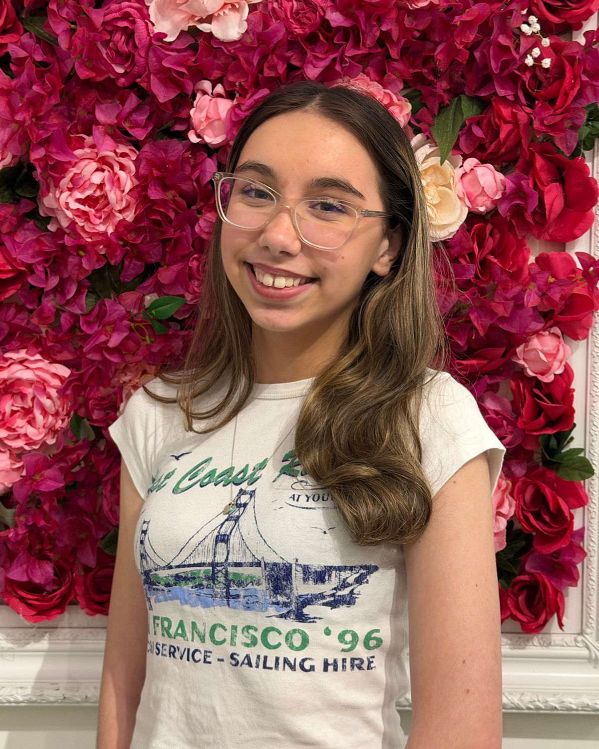 Smiling young woman wearing glasses and a San Francisco 96 t-shirt posed in front of a pink floral wall backdrop.
