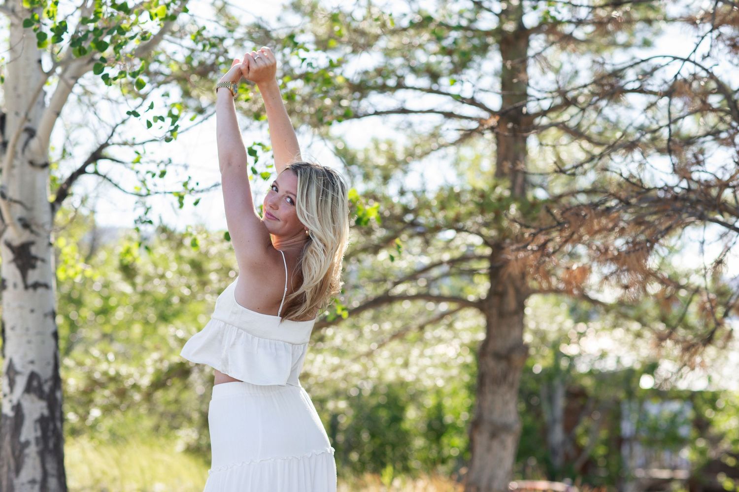 Blonde woman in white off-shoulder dress stretching arms up joyfully surrounded by trees in sunlight.