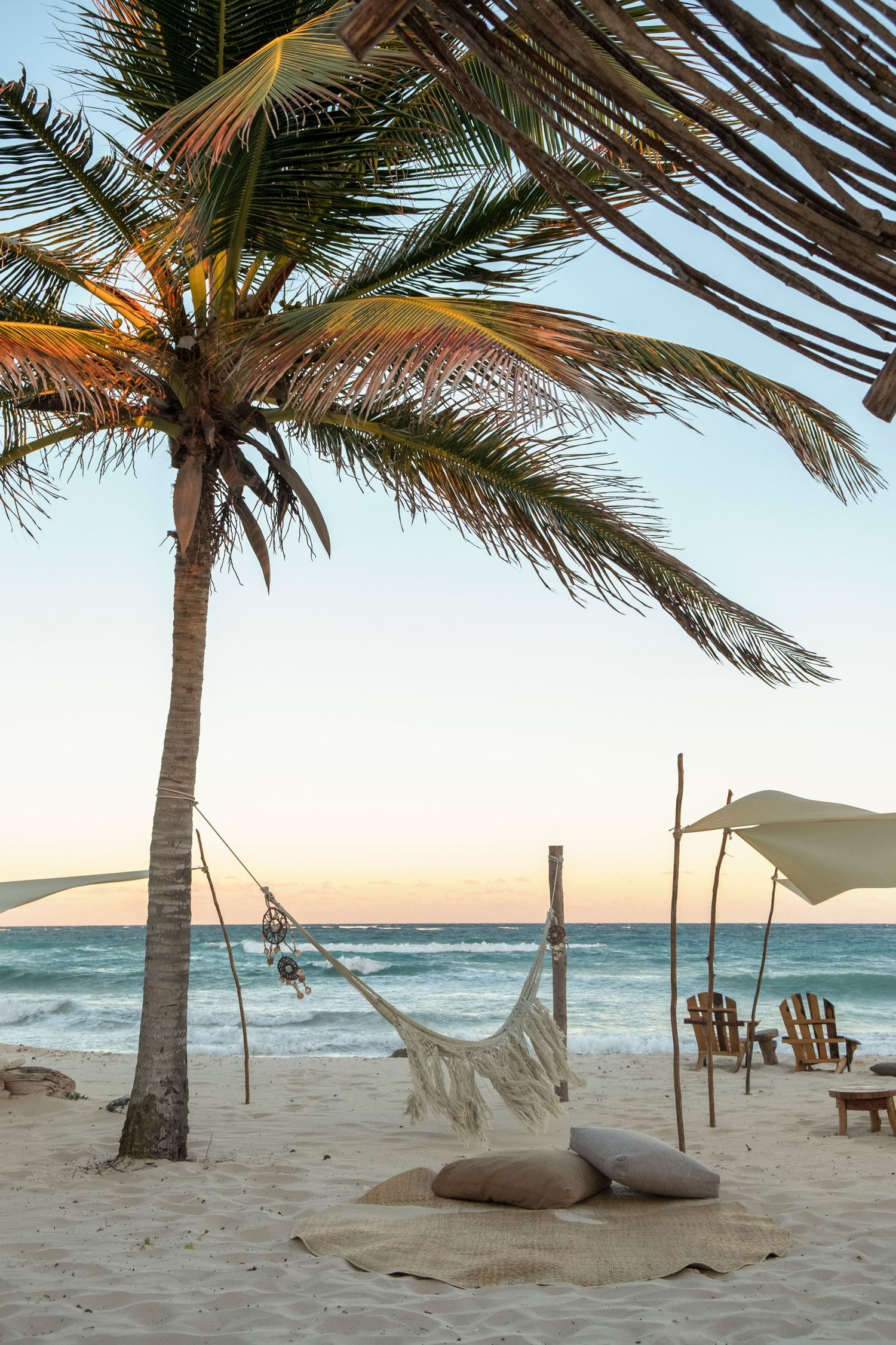 Tropical beach at sunset with a white hammock, palm tree, cushions on sand, and lounge chairs near the ocean.