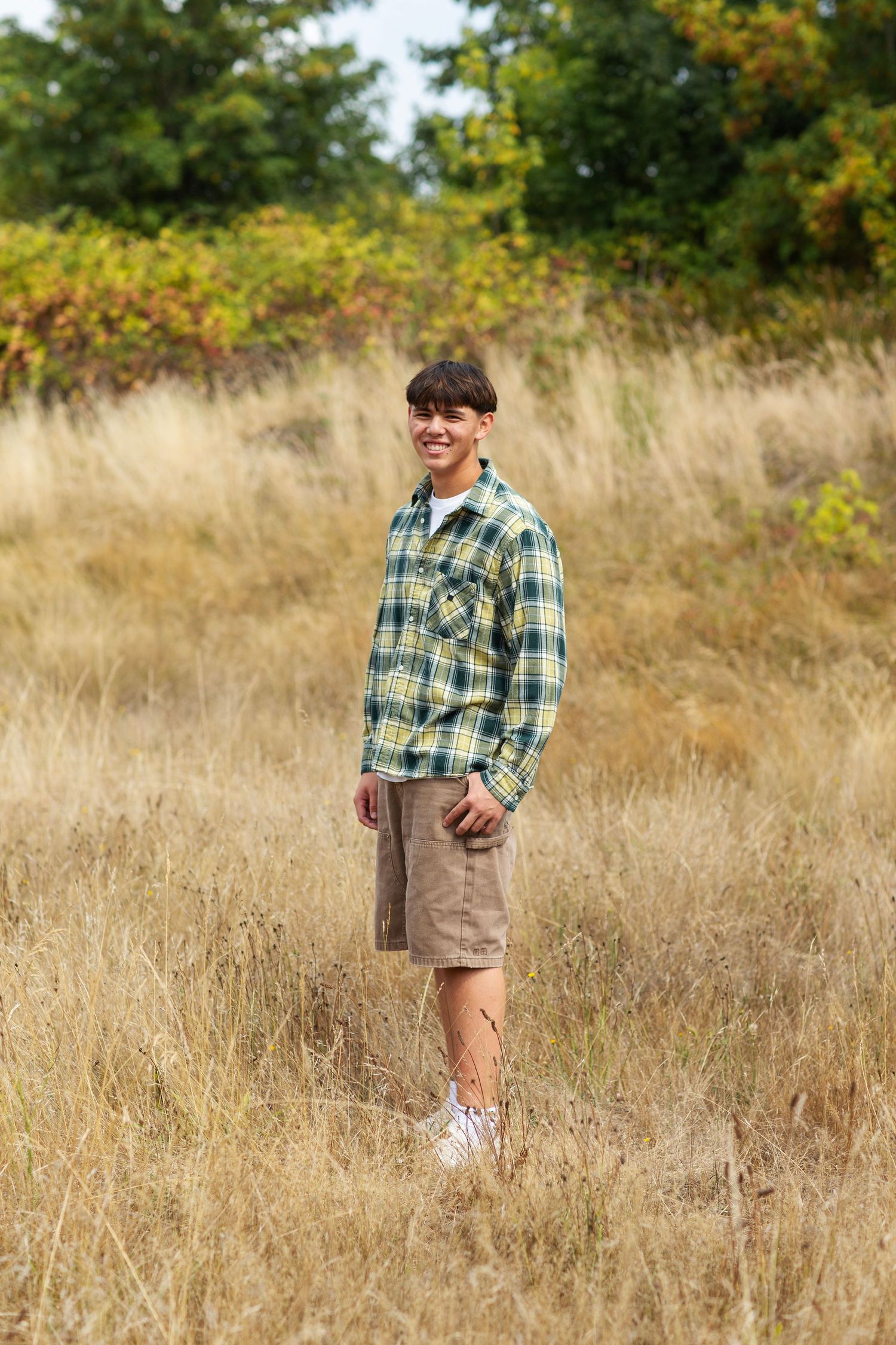 Smiling teen in green plaid shirt and cargo shorts standing in dry grass field.