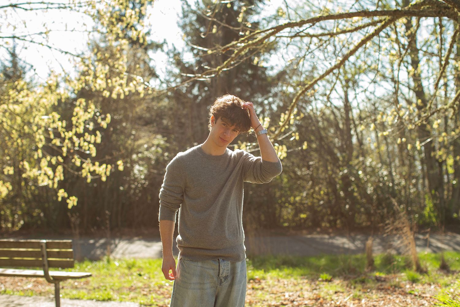 Young man in gray sweater standing in sunny park, hand on head, surrounded by trees and greenery.