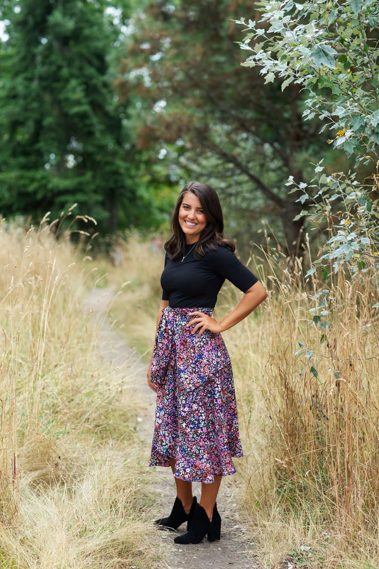 Young woman in black top and floral midi skirt standing on a scenic nature path with tall grass.