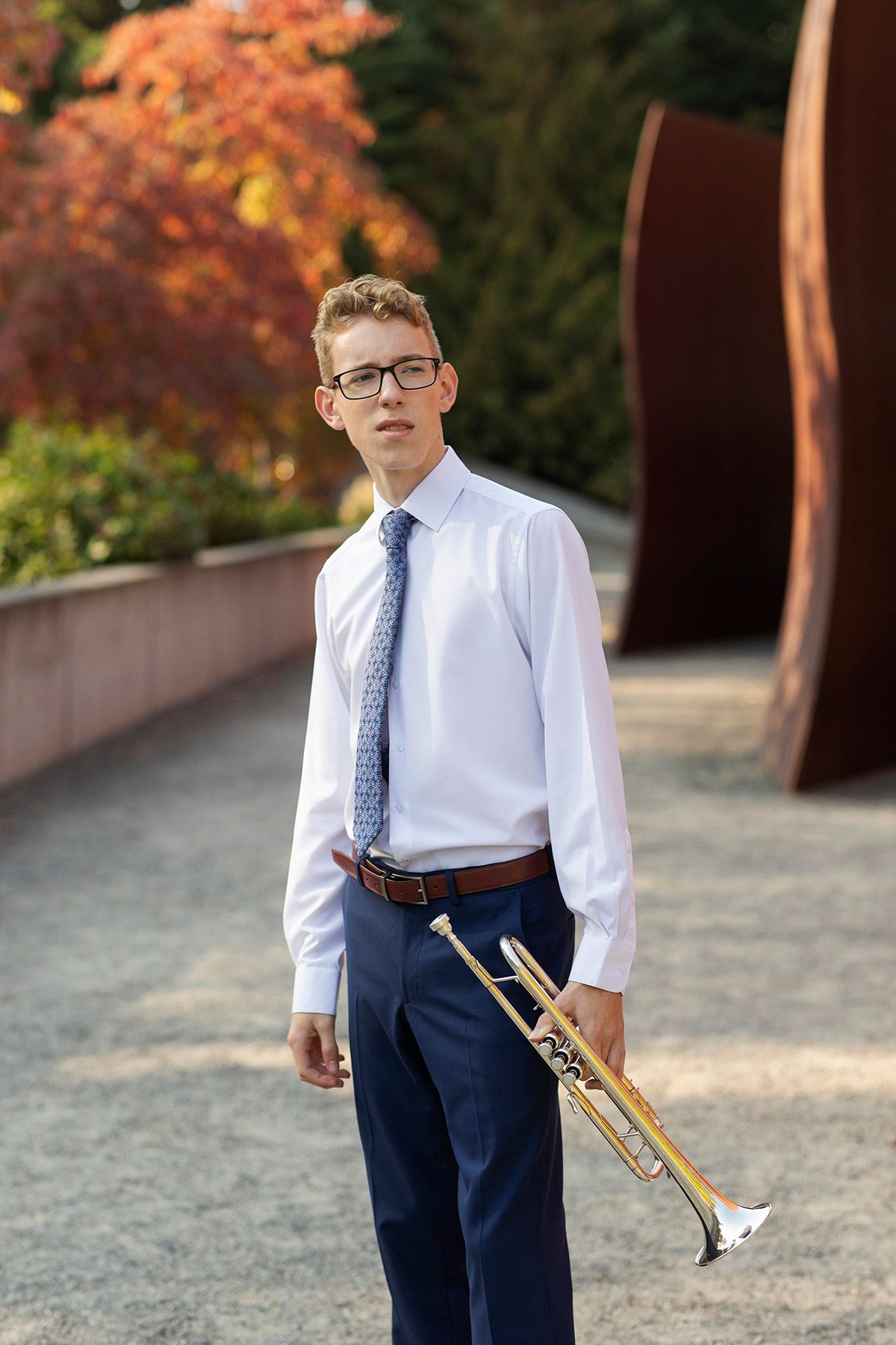 Young musician in white shirt and tie holds trumpet outdoors amid autumn foliage and sculpture.
