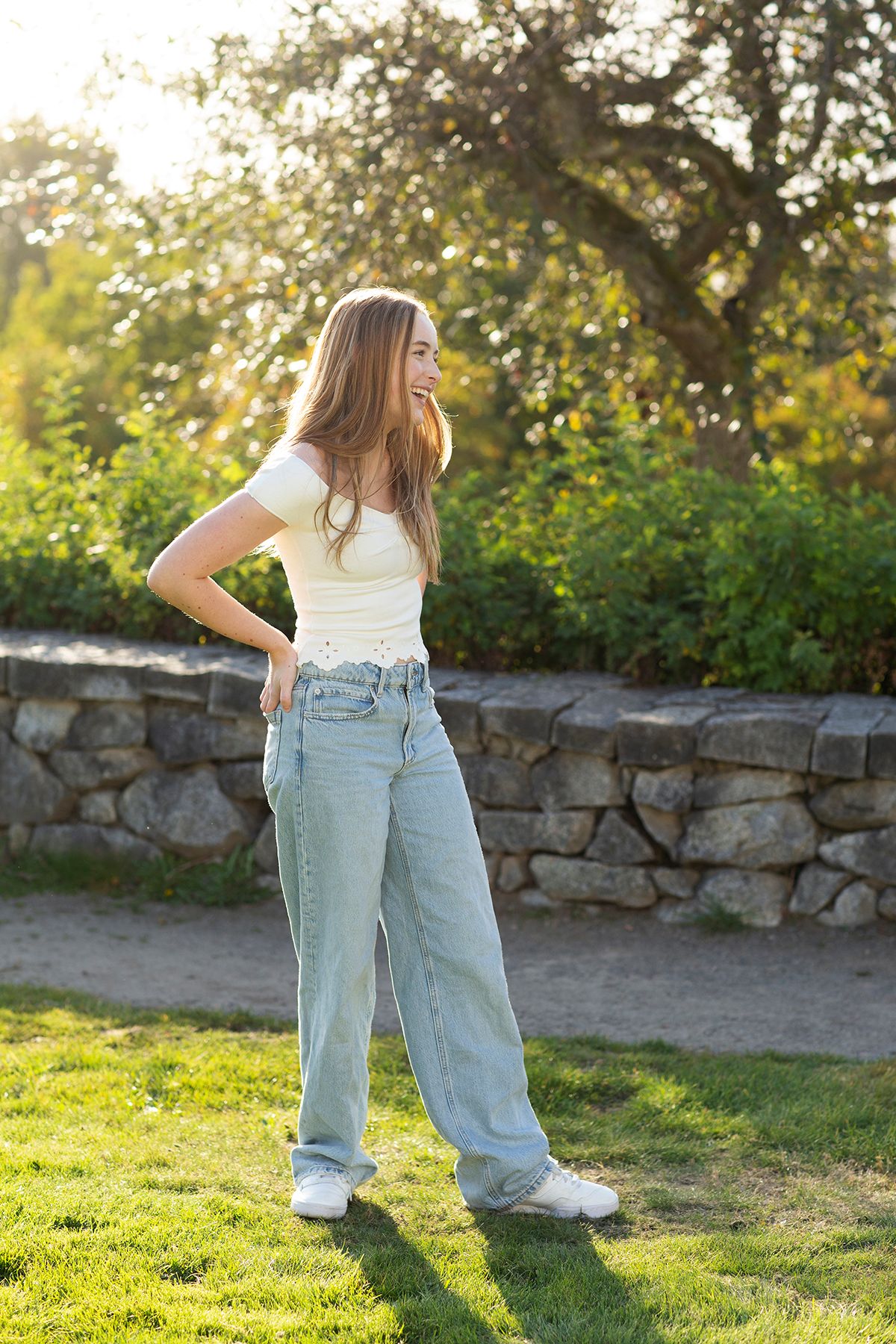 Young woman in white crop top and wide-leg jeans standing outdoors in sunlit park with stone wall background.