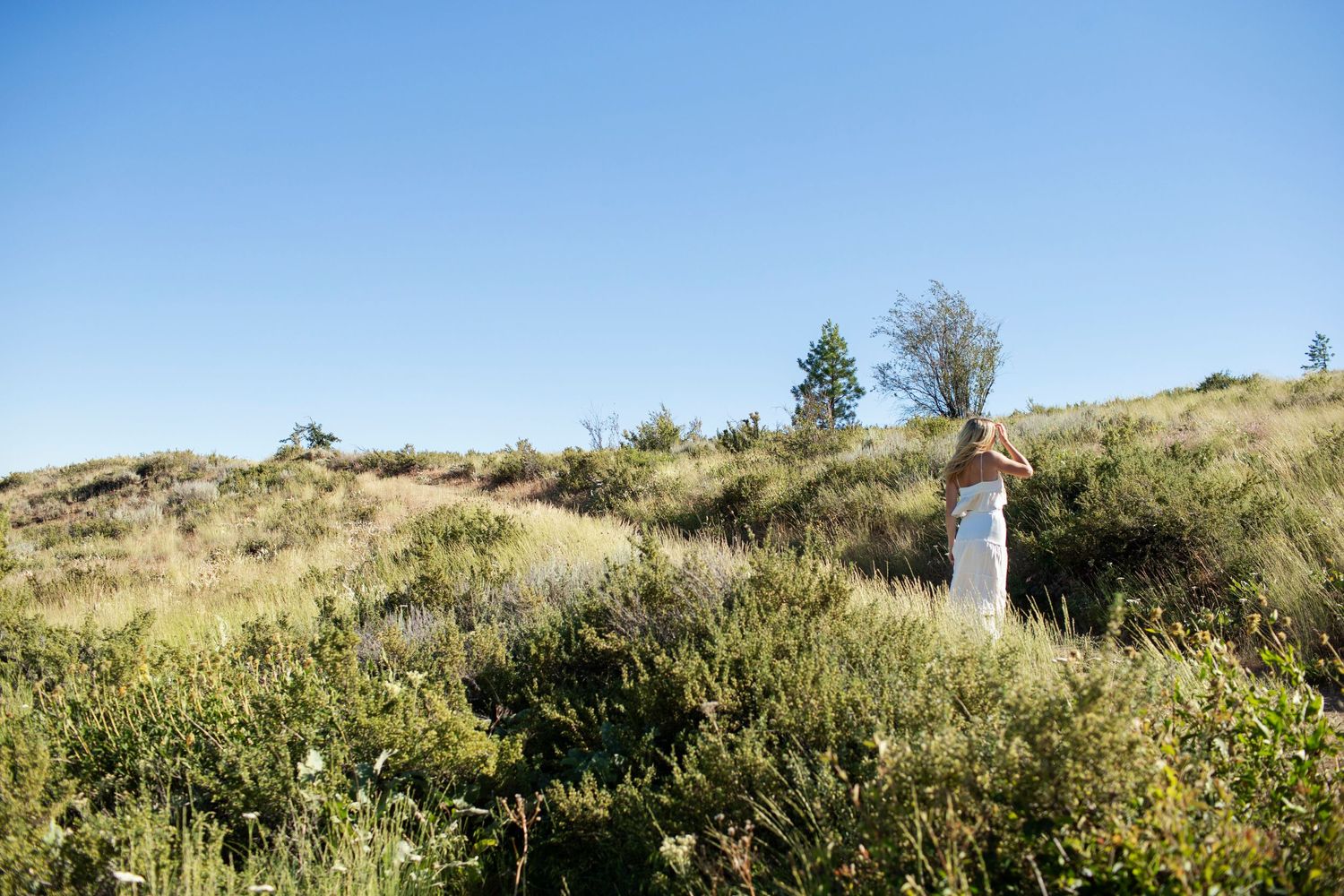 Woman in white dress standing on a grassy hillside under a clear blue sky in nature.