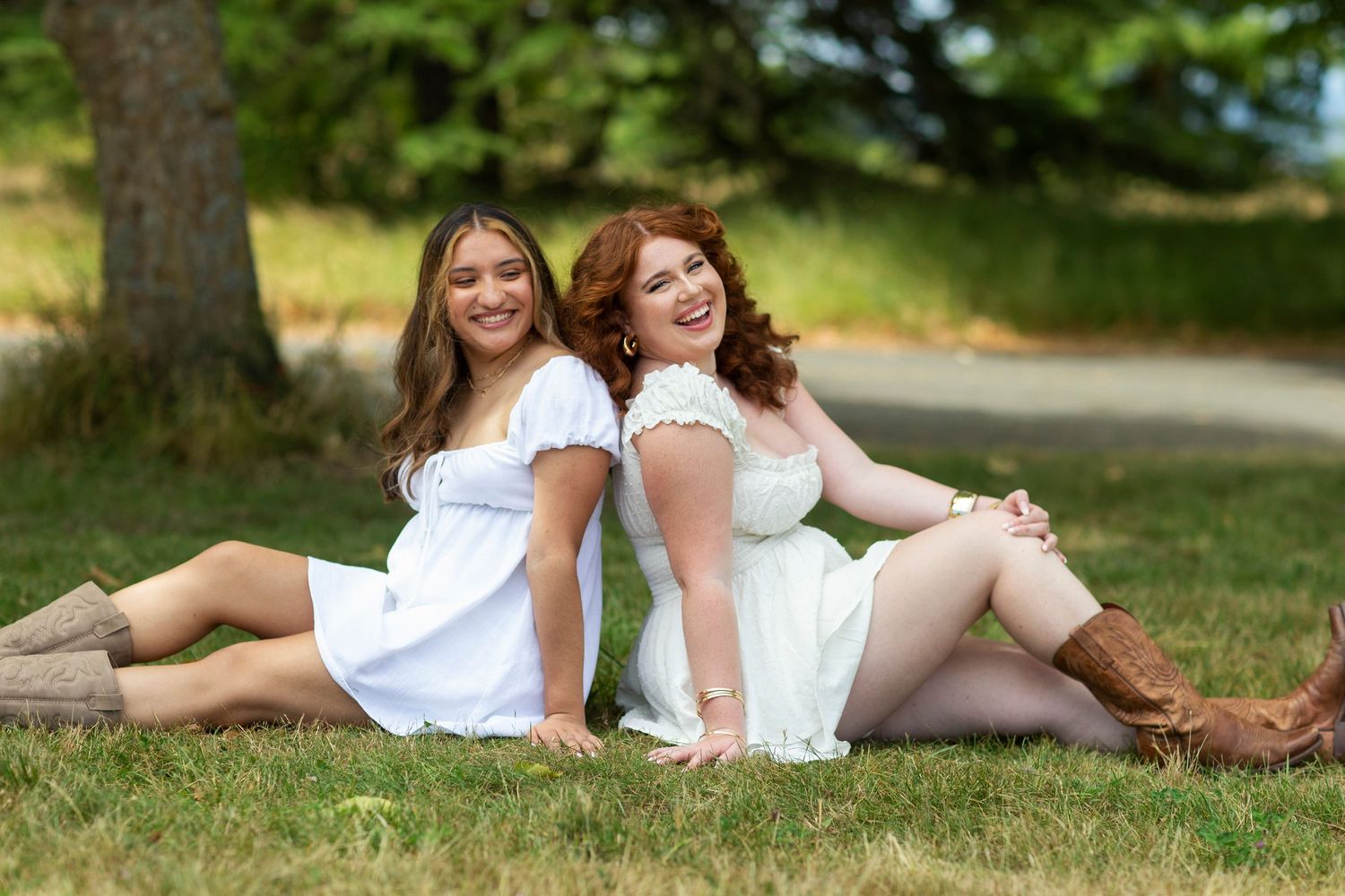 Two young women in white dresses sitting back-to-back on grass.