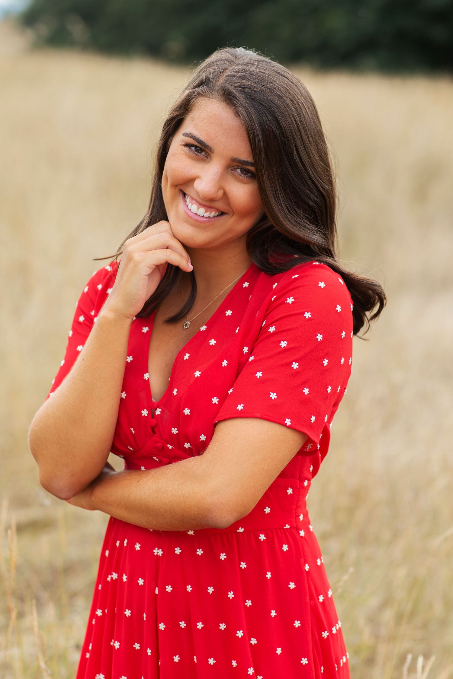 Smiling brunette woman in red floral dress posing outdoors in golden field, hand raised to chin.
