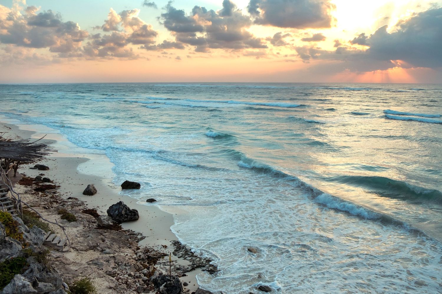 Rocky shoreline with turquoise waves at sunset, featuring a colorful sky and calm beach scenery.