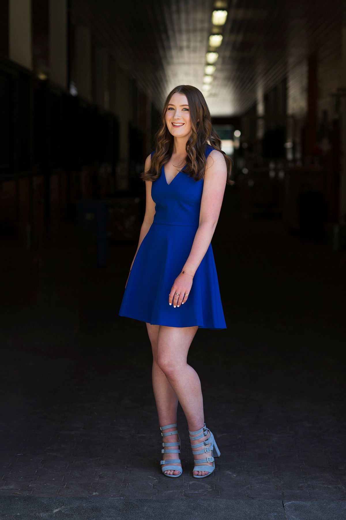 Young woman in royal blue fit-and-flare dress and strappy heels poses confidently in a dramatic industrial hallway.