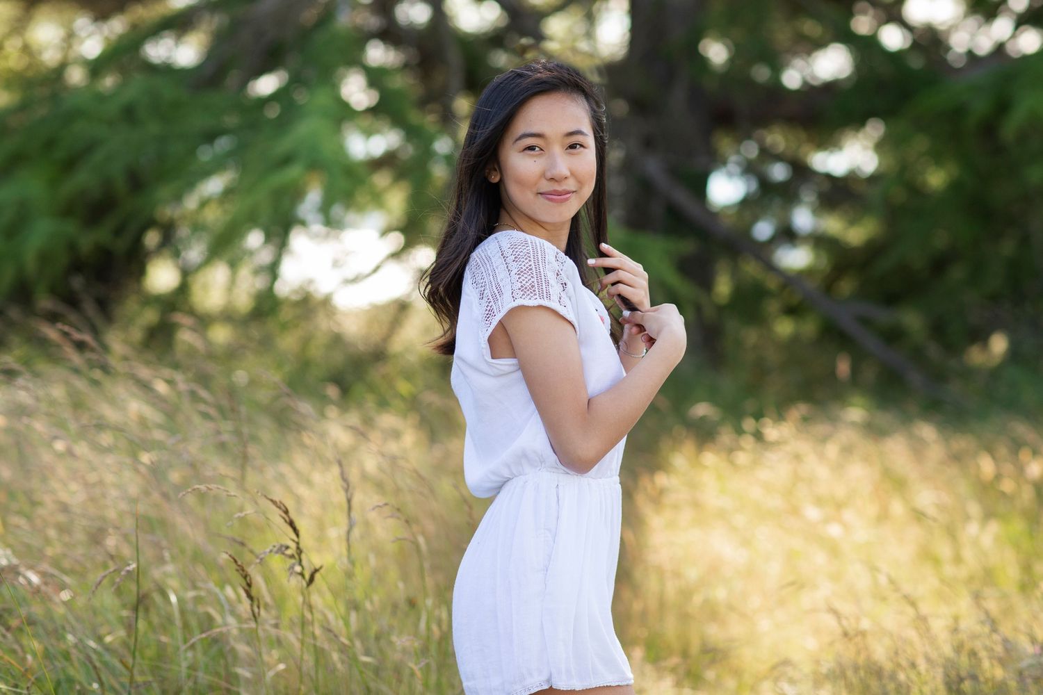 Young Asian woman in white dress standing in golden field with green trees, smiling over shoulder in natural outdoor setting.