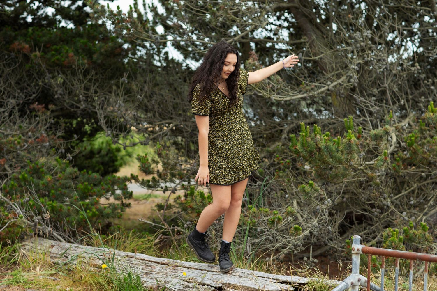 Teen girl in green floral dress balancing on fence rail among coastal shrubs.