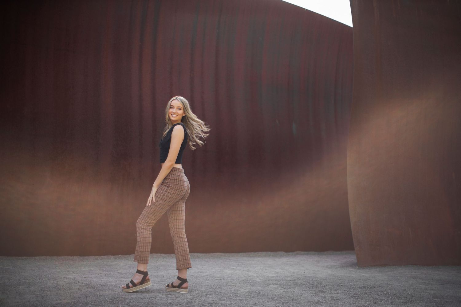 Young woman in plaid pants and black top posing near large rusty metal walls.