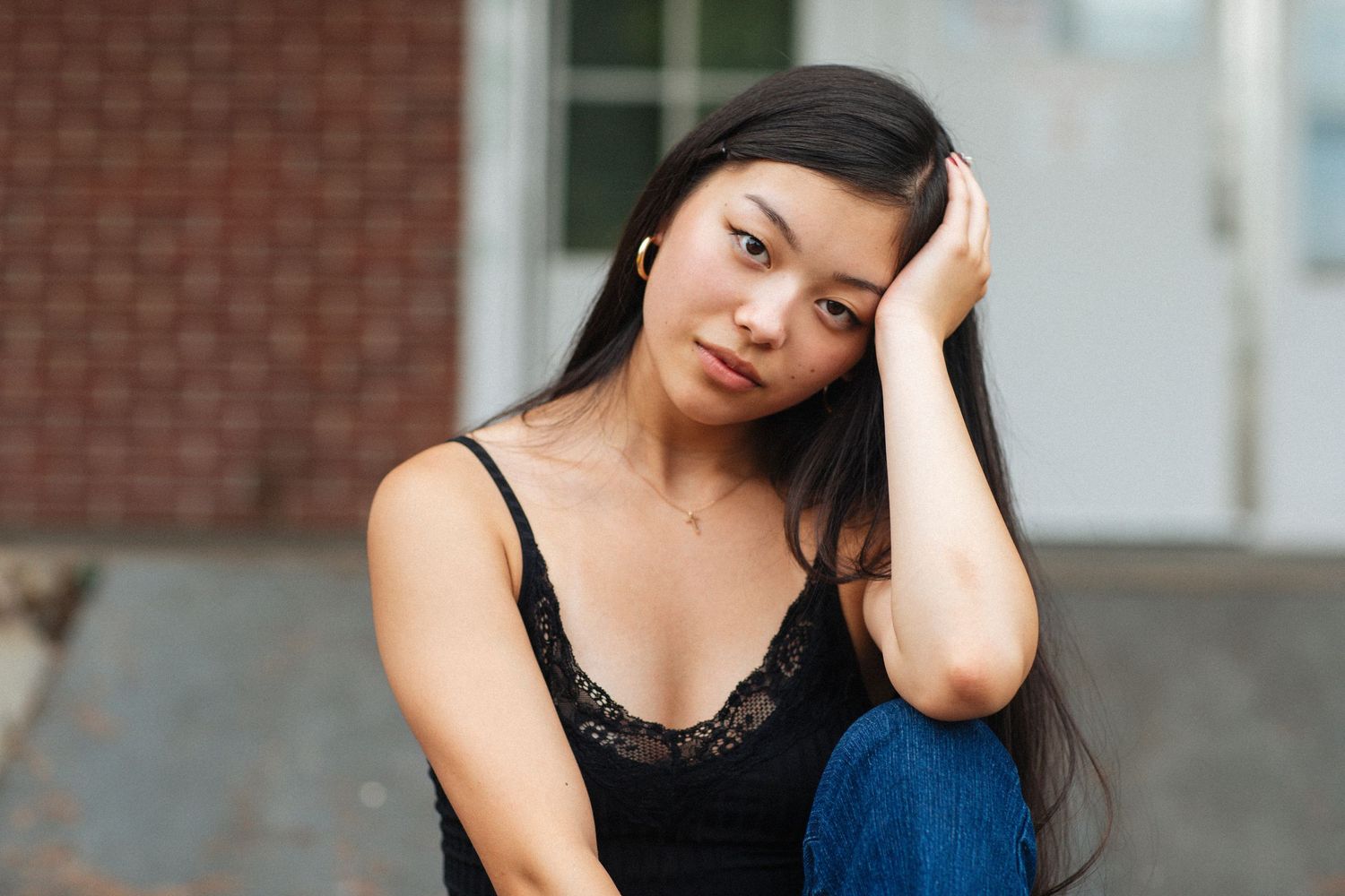 Young woman with long dark hair resting hand on head sitting outdoors near brick building.