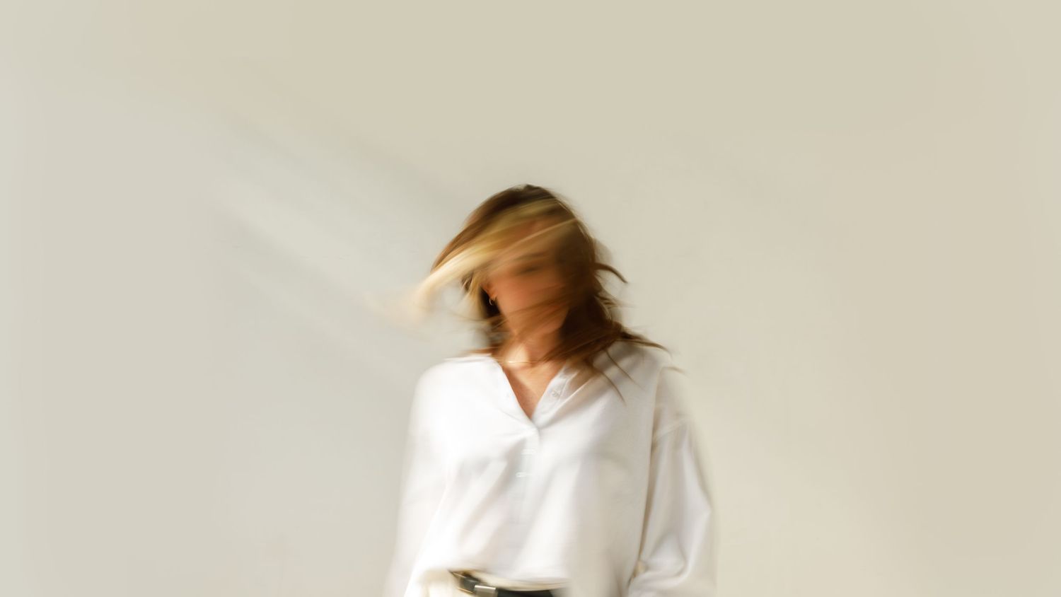 Artistic motion blur portrait of a woman in a white shirt with flowing blonde hair against white background.