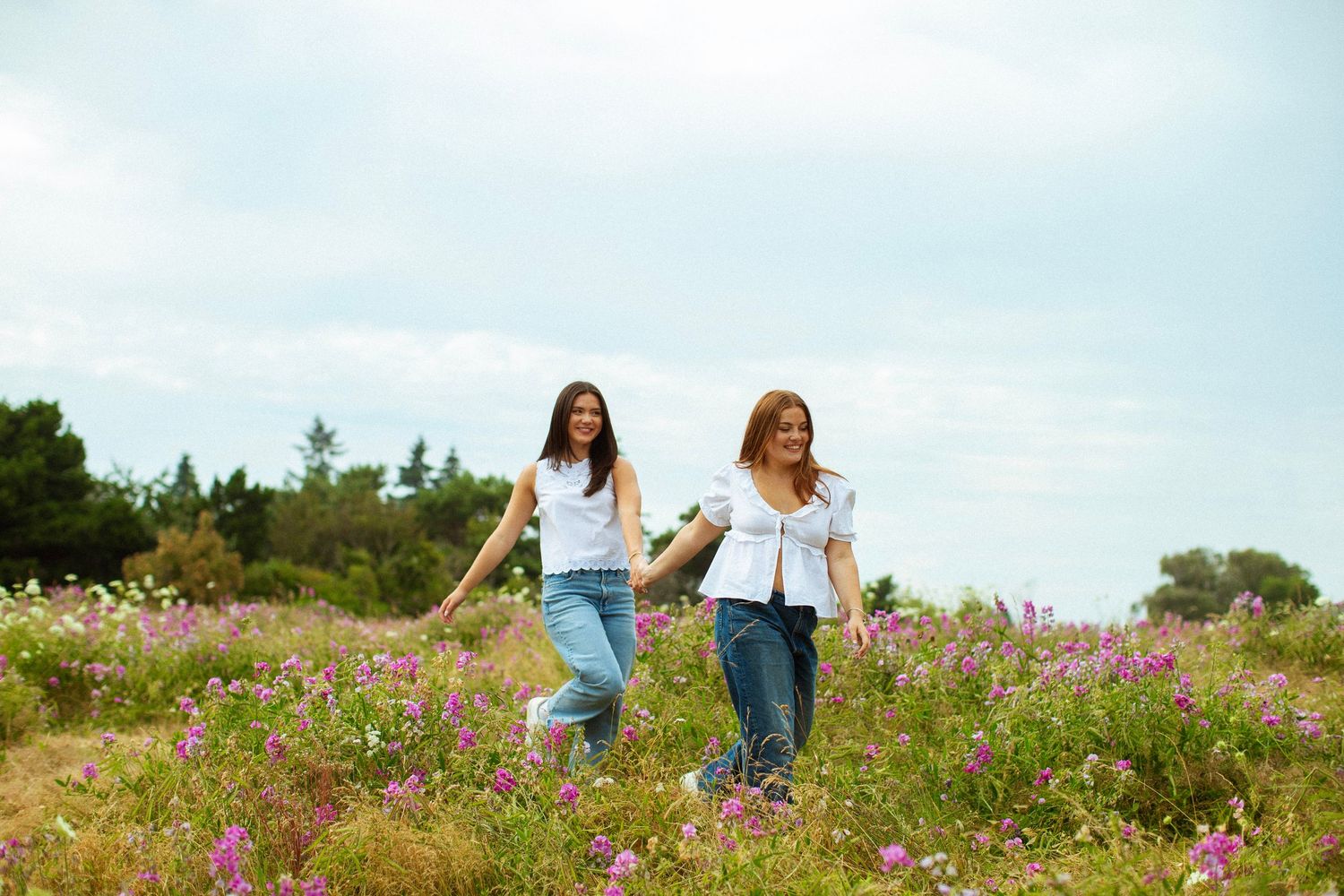 Two women in white tops and jeans walk hand in hand through a vibrant wildflower meadow under a cloudy sky.