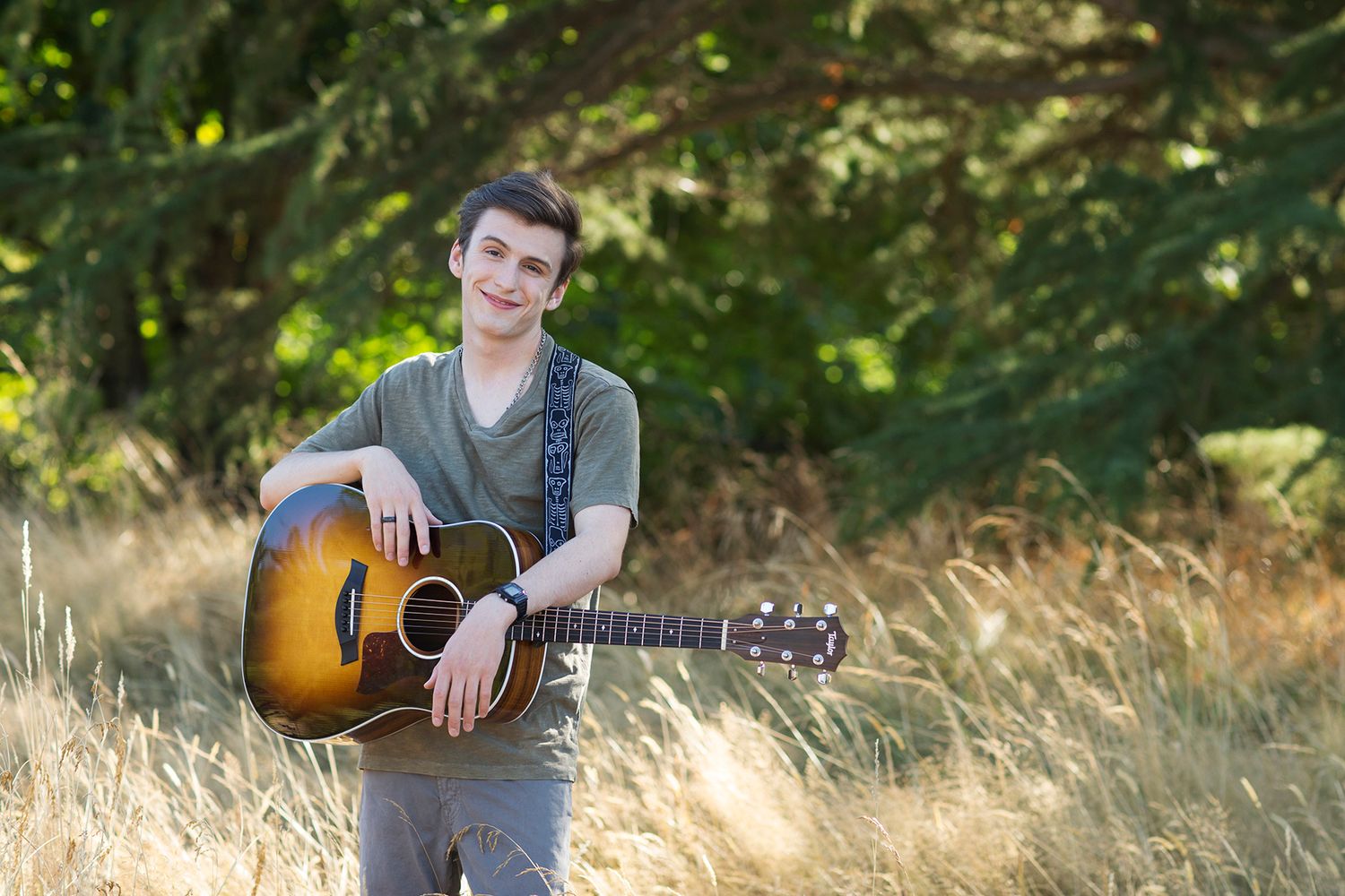 Young man holding an acoustic guitar in a golden field with green trees in the background.