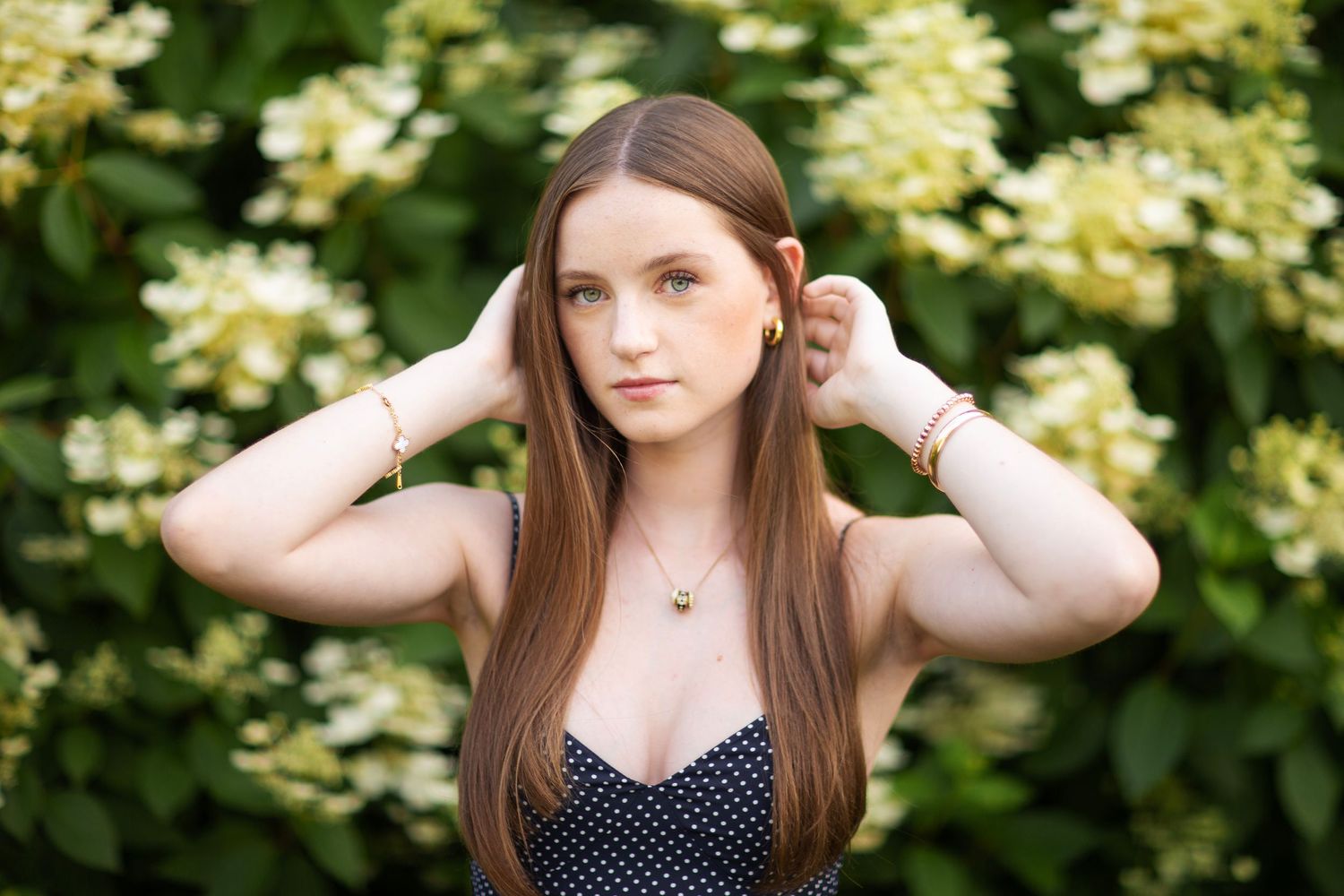 Young woman with long auburn hair poses outdoors among white hydrangea blooms wearing a polka dot top and gold jewelry.