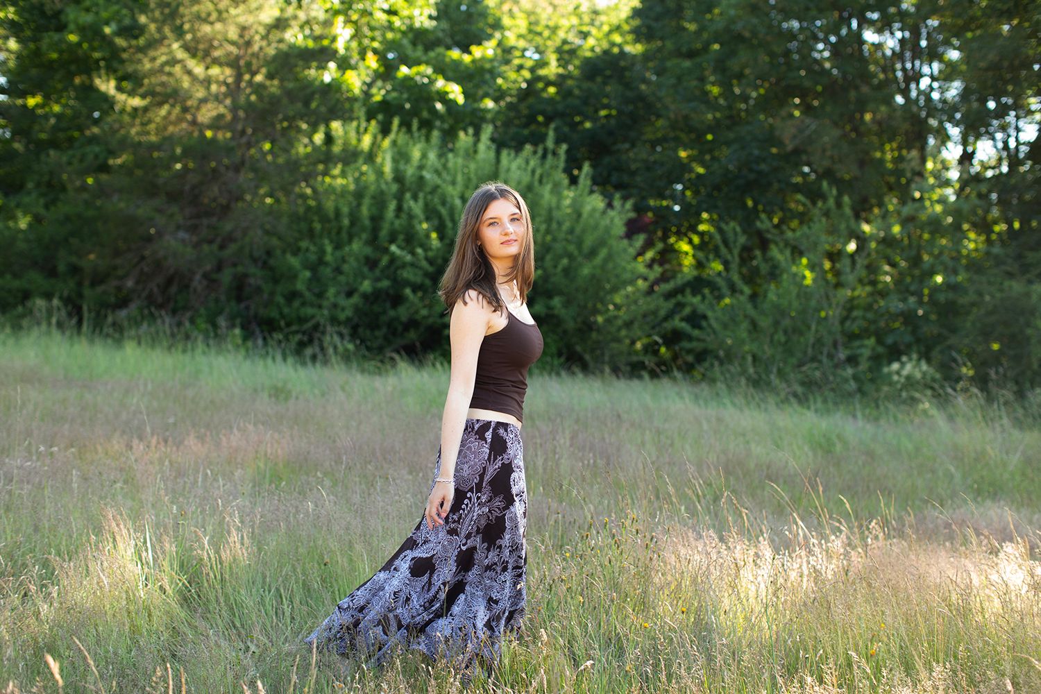 Young woman in brown top and floral maxi skirt standing in a green meadow with trees in background.