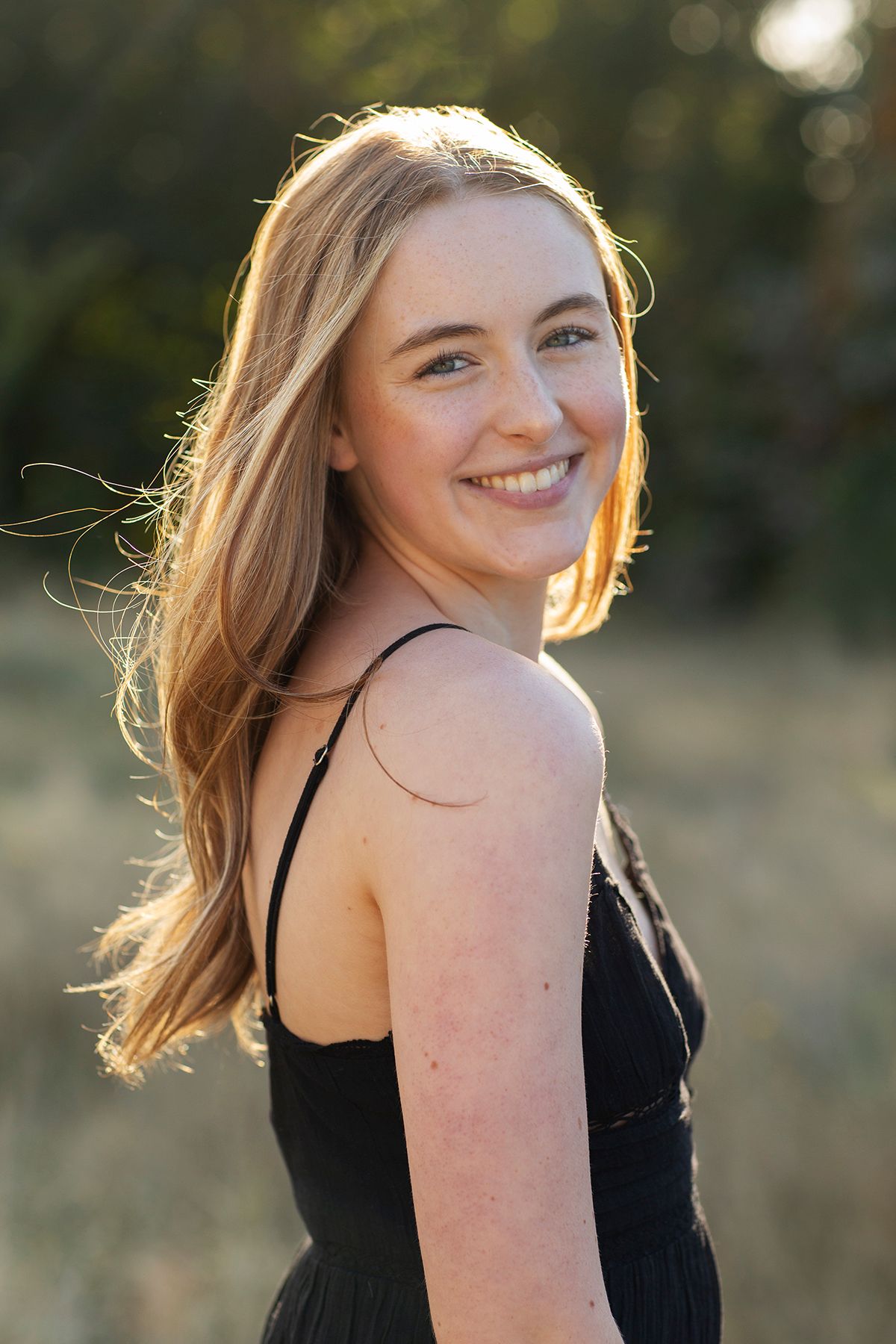 Smiling teenage girl with long blonde hair wearing a black sundress, photographed outdoors in golden hour sunlight.