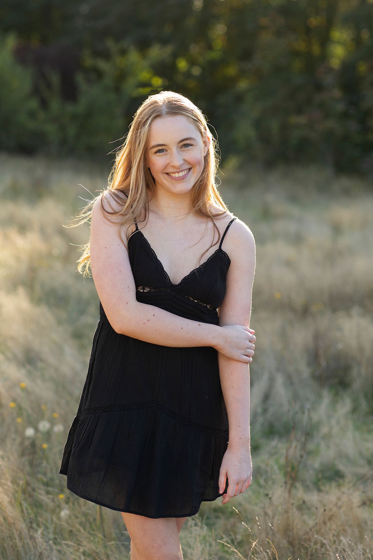 Young blonde woman smiling in a black sundress, standing in a sunlit field with green trees in the background.