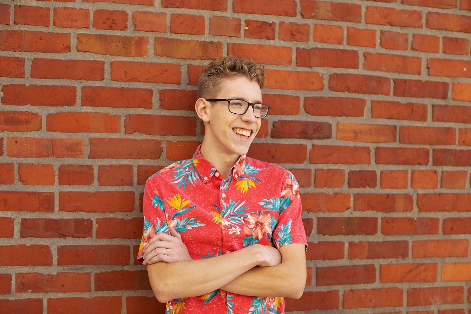 Young man in red floral shirt laughing with arms crossed against a brick wall.