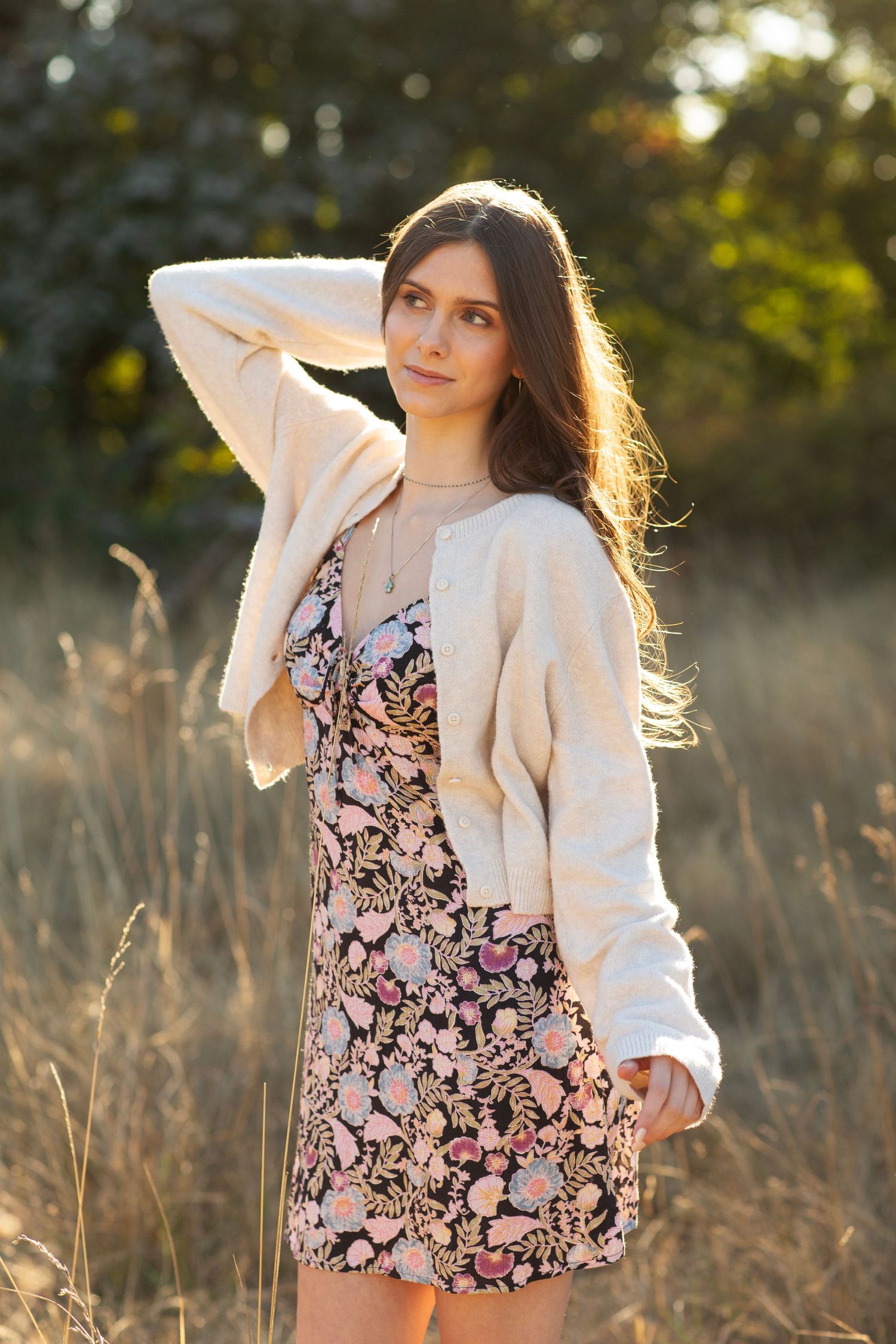 Young woman in floral dress and cream cardigan posing in a golden sunlit field at sunset.
