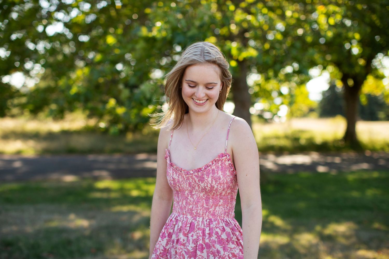 Young woman in pink floral sundress smiling outdoors among green trees.