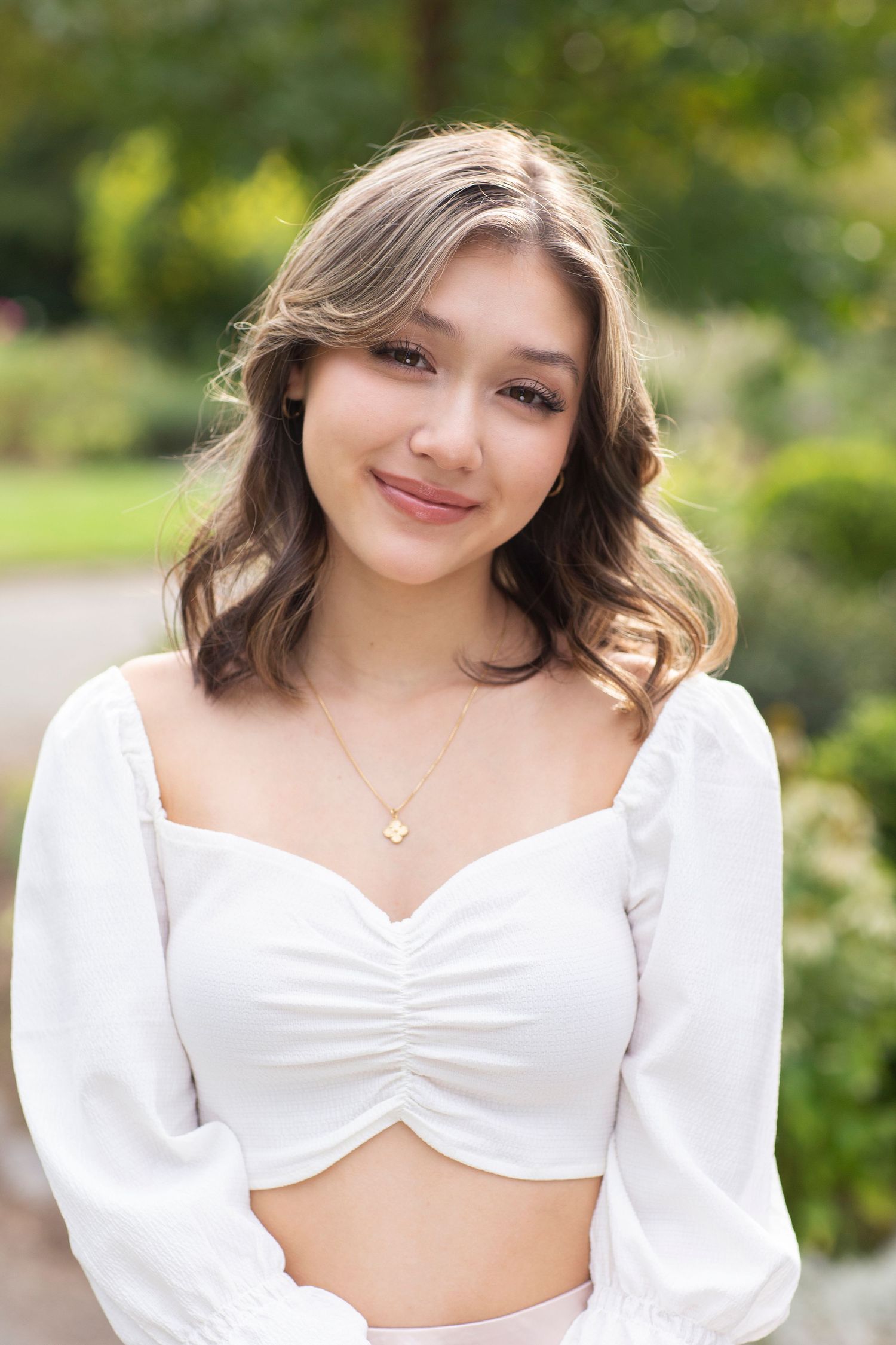Young woman in white crop top with gold necklace smiling outdoors in a lush green garden setting.