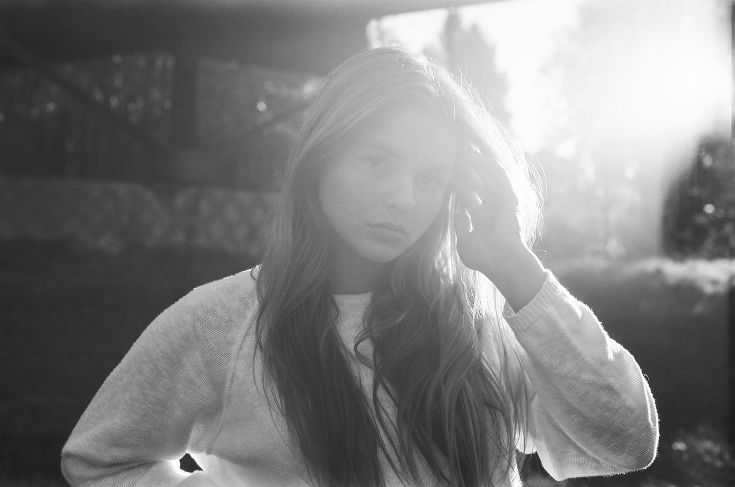 Black and white portrait of a young woman with long hair, hand raised to her head, soft backlit sunlight.