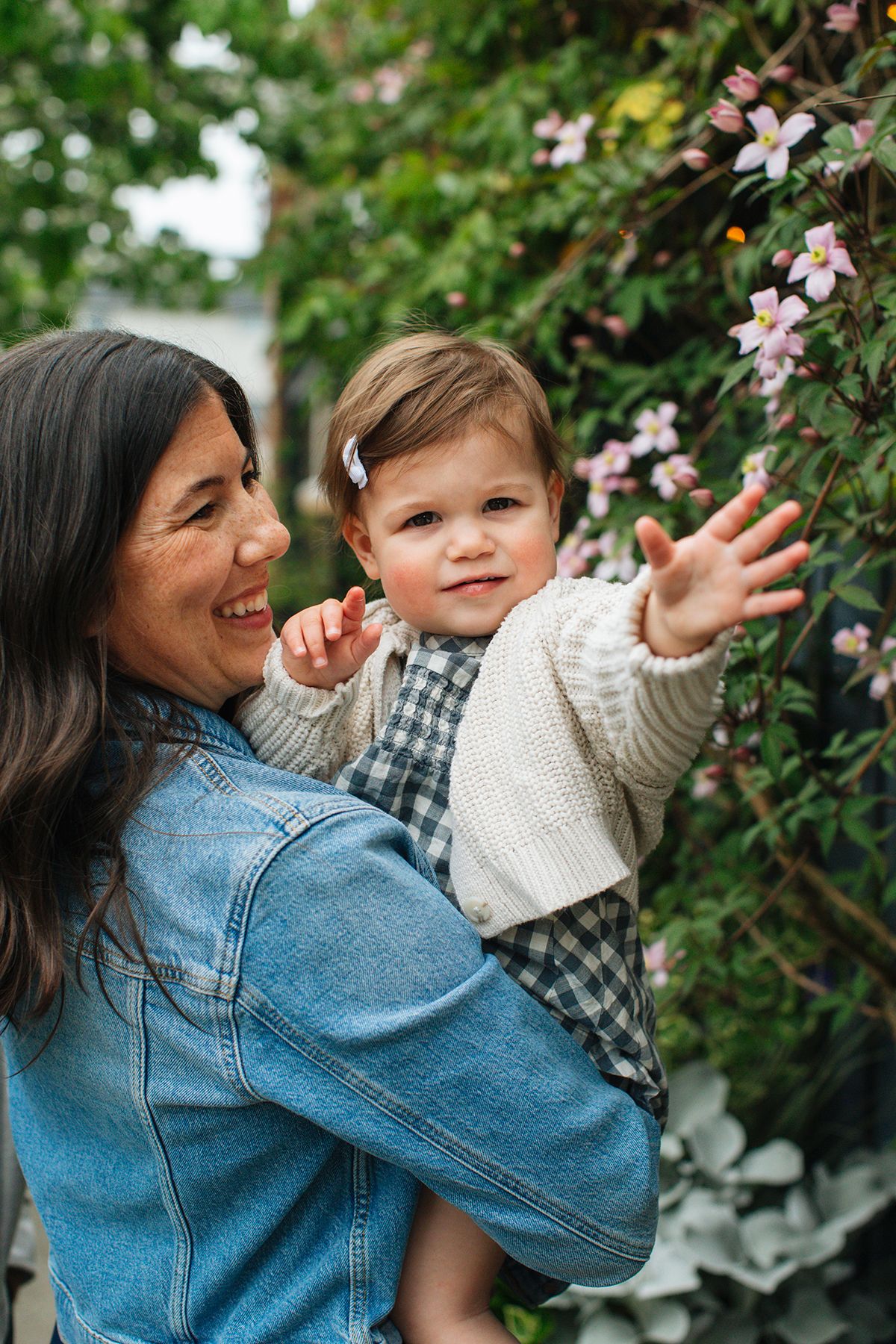 A smiling woman in a denim jacket holds a toddler girl reaching toward pink flowers in a lush garden.
